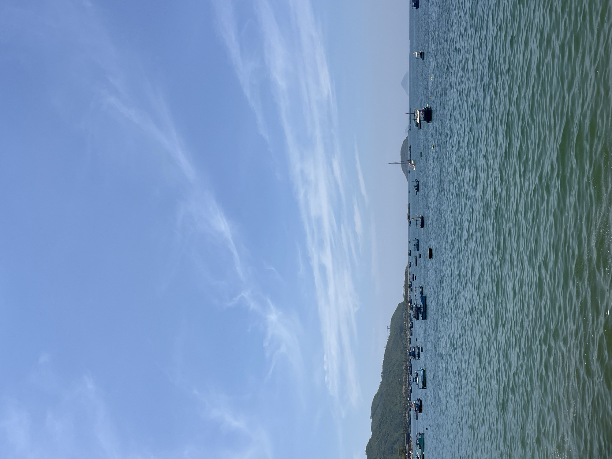 This image displays a coastal scene in Bac Nha Trang Ward, Vietnam. The foreground consists of rippled, blue-green ocean water. Numerous small fishing boats, primarily dark blue and brown-hulled, are dispersed across the water, appearing to be anchored or moored. One white-masted sailboat is also present among the vessels, along with several yellow marker buoys. In the background, a large, verdant mountain range covered in trees extends along the coastline on the left, with visible signs of development or construction, including a crane, near its peak. To the right, two hazy, conical landmasses or islands are discernible on the distant horizon. The upper portion of the image reveals a clear, light blue sky with scattered, thin, white wispy clouds. No individuals are visible within the frame.