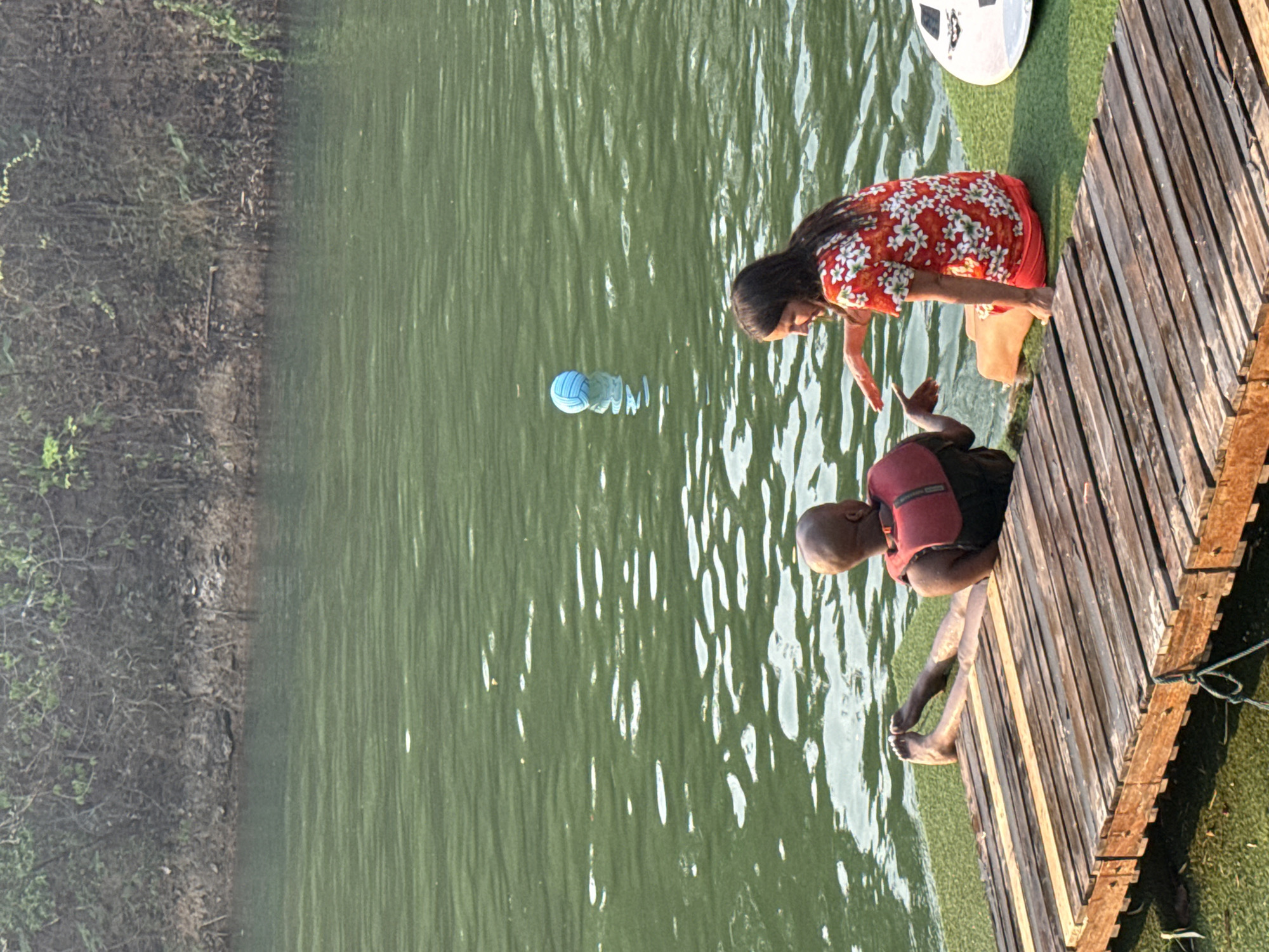 A woman and a man are seen on a wooden pier extending into a body of water. The woman, wearing a red floral dress, is reaching towards the man. The man is sitting on the edge of the pier, wearing a red life jacket, with his feet dangling in the water. A blue volleyball is floating in the water near them. The pier is constructed of wooden planks. The water appears greenish. Vegetation is visible on the shoreline opposite the pier. The scene is within Tambon Nam Phrae, Thailand.