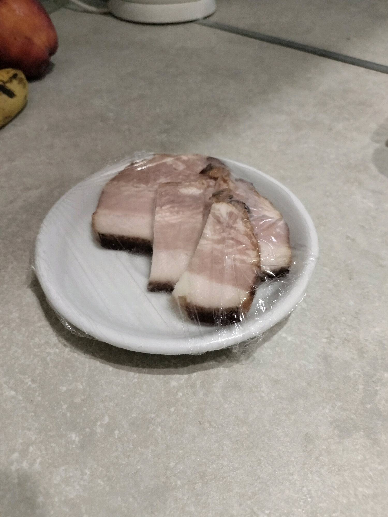 A white plate covered in plastic wrap contains four slices of cured meat with visible fat and lean layers. The plate is on a textured, light gray surface, possibly a kitchen countertop. In the background and to the left, parts of a red fruit and a banana are visible. The scene is indoors, likely in a residential kitchen.