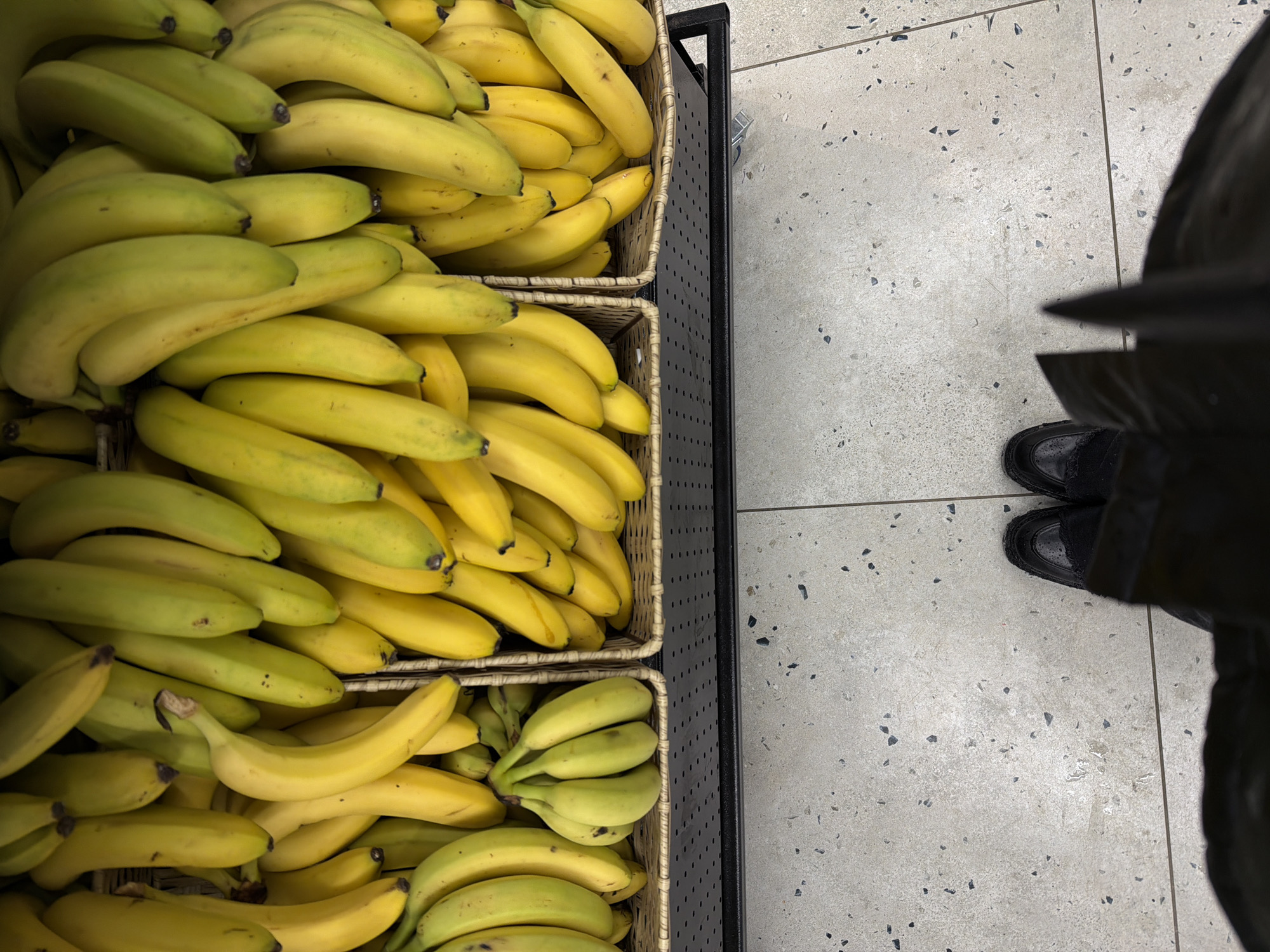 The image shows a selection of bananas displayed in a three-tiered wicker basket, situated next to a person. The bananas range in ripeness, from slightly green to fully yellow. The basket is part of a black metal display stand. The person, partially visible on the right side of the frame, is wearing black shoes. The setting appears to be indoors, with light gray tile flooring. The image's location is not discernible as Moscow, Russia, from the visual elements provided.