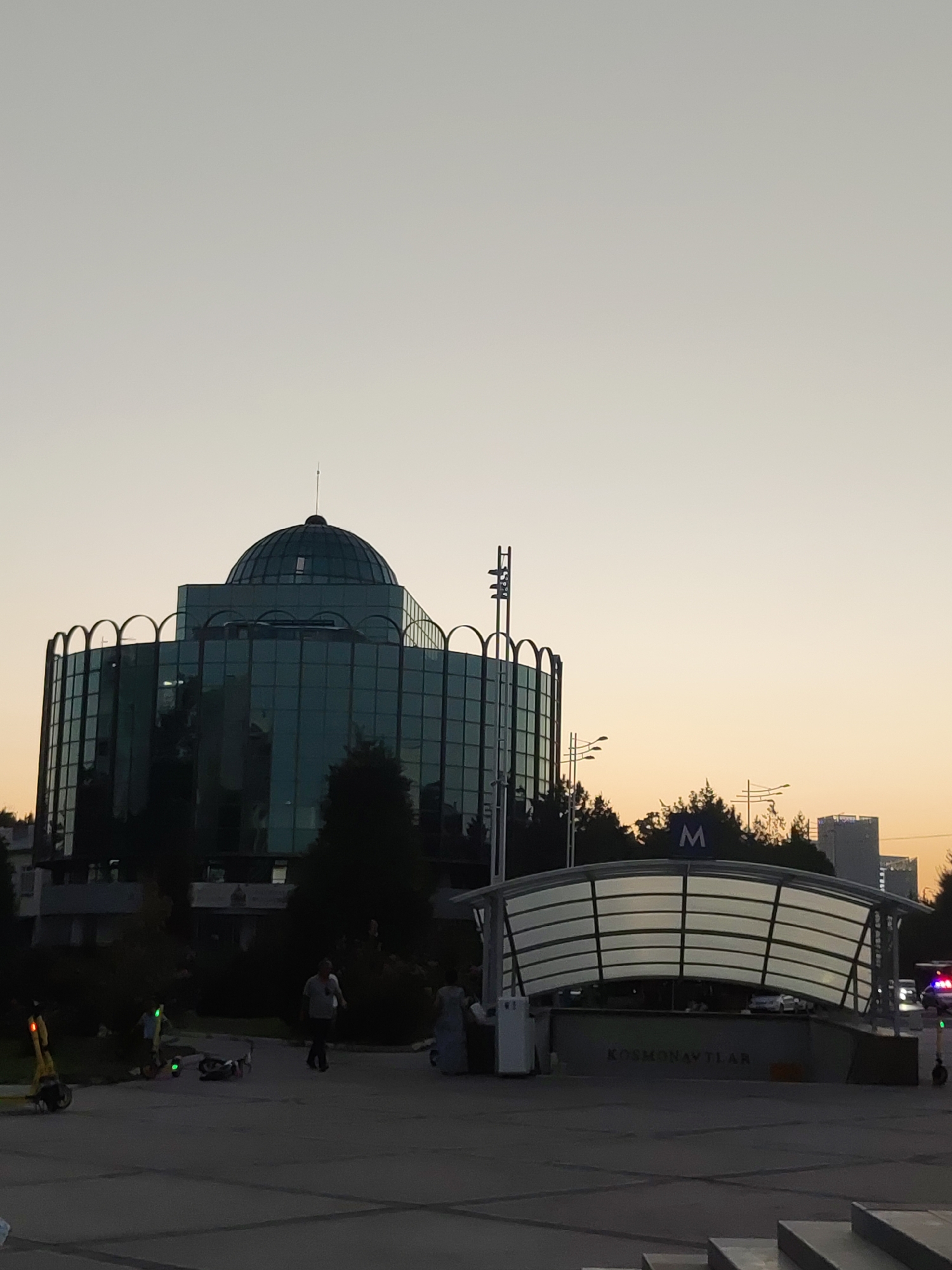 The image depicts the Kosmonavtlar metro station in Tashkent, Uzbekistan, at dusk. A large, modern building with a glass facade and a dome structure is visible in the background. In the foreground, the arched entrance of the Kosmonavtlar metro station is illuminated, with the name of the station written on its facade. A blue sign with a white "M" indicating the metro is positioned above the entrance. Several people are walking in the plaza area, and a yellow electric scooter is parked to the left. Trees and distant city buildings are also present. The sky is a pale orange, suggesting the end of daylight.