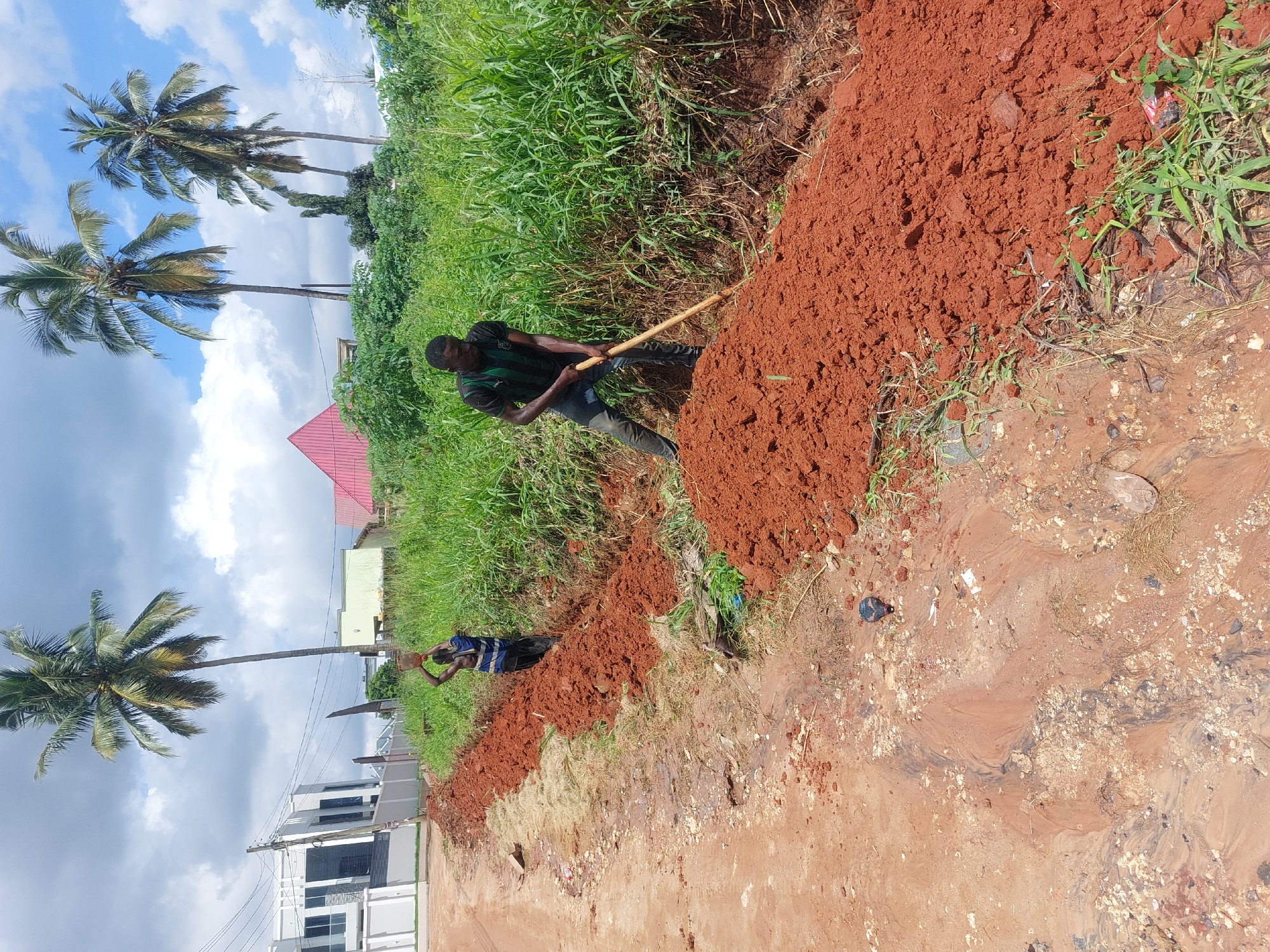 The image captures a scene of manual labor in Dar es-Salaam, Tanzania. Two men are working on leveling red soil along a roadside. One man is wielding a shovel, redistributing the soil, while the other is positioned further down the stretch of soil, possibly also involved in the same task. Lush green vegetation and palm trees are visible in the background, indicative of the tropical environment. Buildings are scattered in the distance, suggesting an urban or peri-urban setting. The road appears to be unpaved and has evidence of water flow, creating a muddy texture. The sky is partly cloudy, with patches of blue visible.