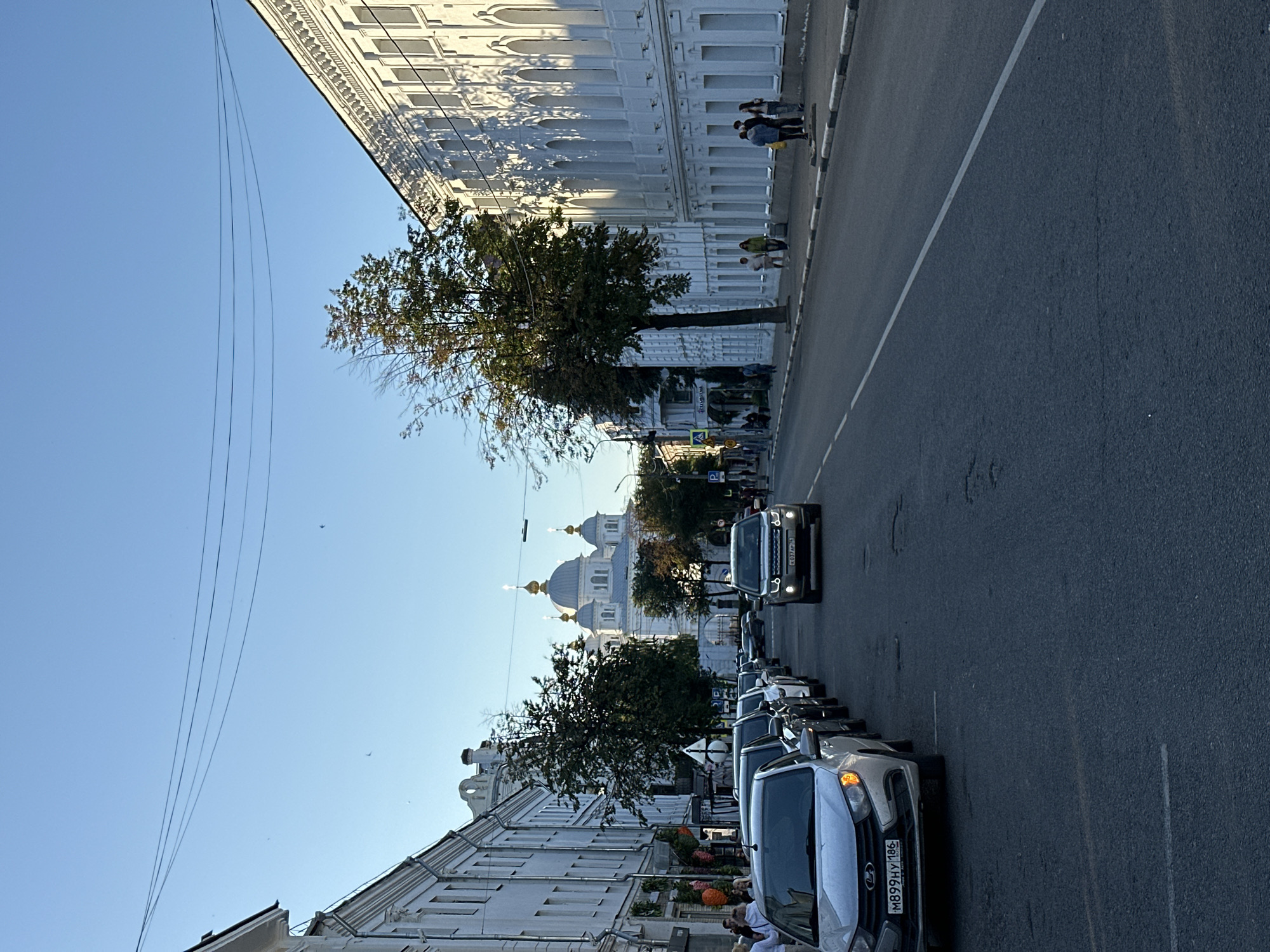 The image depicts a street scene in Yazvitsevo, Russia, under a clear blue sky. On the left side of the street, several white buildings with decorative facades line the road. Some of these buildings have multiple windows and architectural embellishments. A row of cars is parked along the side of the street, with the closest vehicle being a silver sedan. Further down the street, a white SUV is in motion.
On the right side of the street, a long, white building with a highly ornate facade, featuring numerous arched windows, dominates the view. A large tree with green and reddish leaves stands between the building and the road. Several people are visible walking on the sidewalk in front of this building.
In the distance, at the end of the street, a large white church with multiple golden domes is prominently featured. The street itself is asphalt with white lane markings. Power lines are visible in the sky, strung between poles. The overall impression is a daytime street view in a Russian town or city.