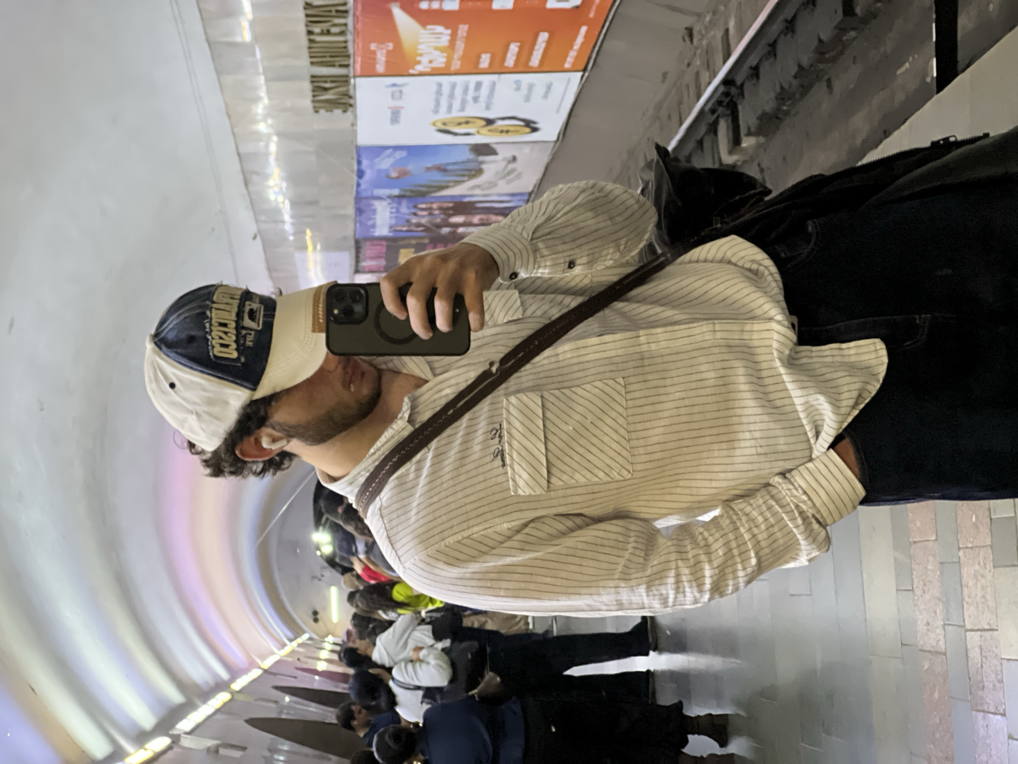 A man in a baseball cap, striped shirt, and dark pants is taking a selfie with a smartphone in the Yerevan metro in Armenia. He has a messenger bag over his shoulder. Behind him are a row of advertisements and a crowd of people moving through the tunnel-like metro station.