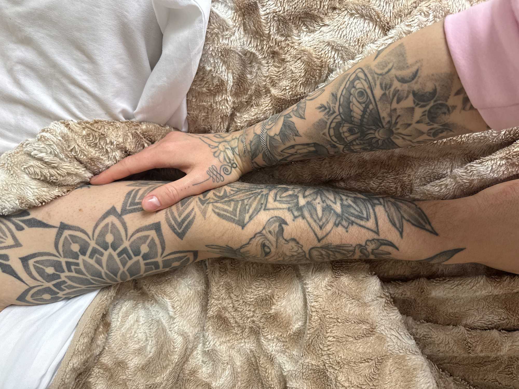 The image shows two tattooed arms resting on a beige, textured blanket. A hand, also tattooed, rests on the left arm. The tattoos are primarily in black ink and depict various designs, including mandalas, a butterfly, and other geometric patterns. The arm closest to the top of the image also has a thin, silver-colored chain bracelet. The left side of the image includes white bedding. The location is not discernable from the provided image.