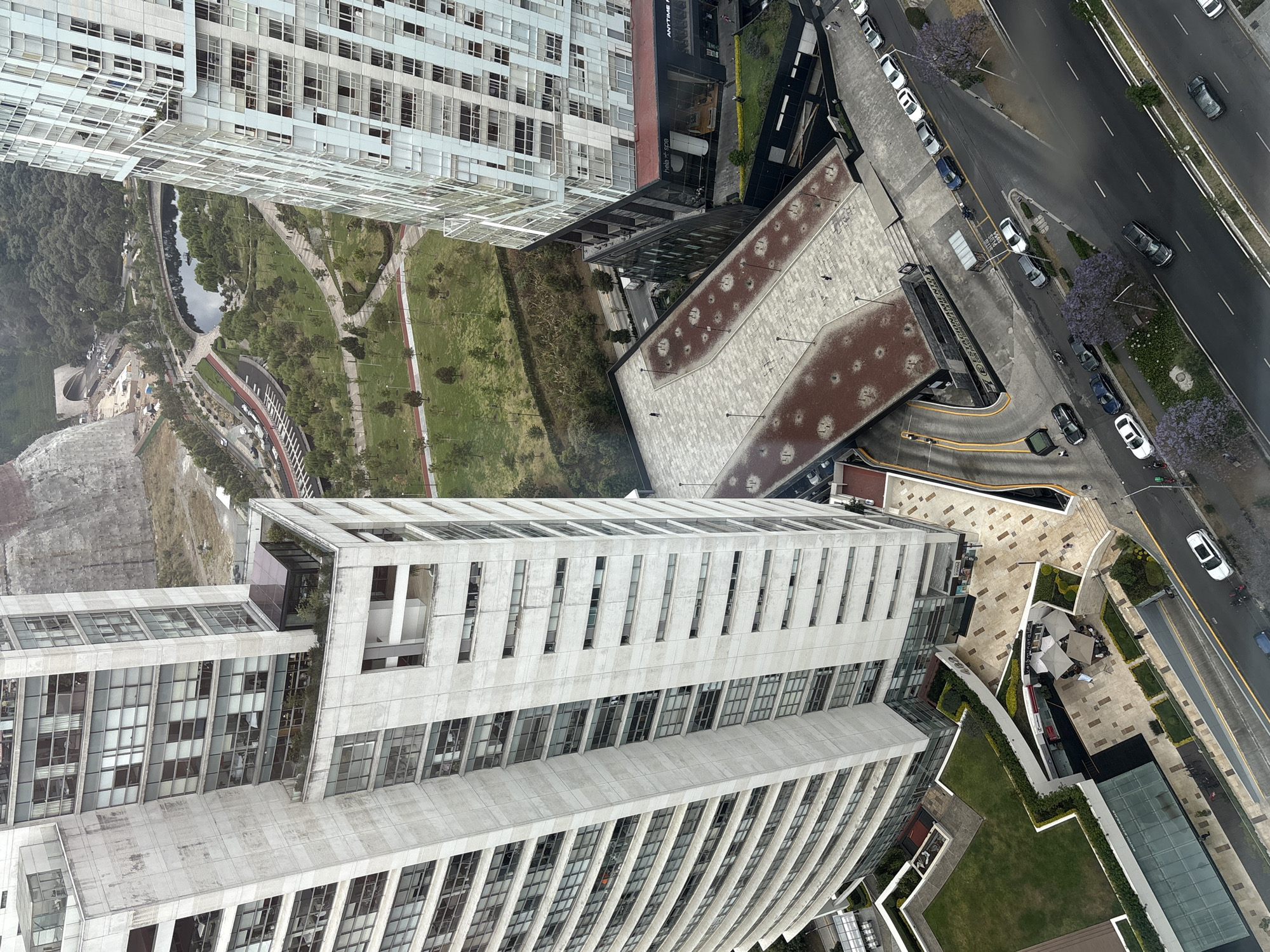 This high-angle, aerial photograph captures a modern urban landscape in Mexico City, featuring several tall buildings, a large park, and a multi-lane road. The image is taken from a significant height, looking down into the city.
In the foreground, a prominent modern building with a concrete facade and numerous rectangular windows stands tall, displaying a unique "wavy" architectural element on its lower levels. Adjacent to it, a large, flat rooftop of another structure is visible, characterized by a grey surface patterned with many reddish-brown circular spots.
A wide, multi-lane road runs across the mid-ground and right side of the image, showing cars driving and parked along the side. Notable purple-flowering Jacaranda trees line the median and sidewalks of the road, suggesting a specific season. Several vehicles are visible, including white, grey, and black cars, and a motorcycle. One black truck has "VAG" visible on its rear.
Beyond the road and structures, a sprawling green park occupies the left and central portion of the scene. It features winding paved paths, vast grassy areas, and numerous trees. A tranquil lake or pond is nestled within the park, reflecting the overcast sky. Further into the park, infrastructure like a running track (red surface) and a tunnel entrance are discernible.
Other buildings in the scene include a tall, white building with a distinct gridded facade of windows and balconies, and a structure with a glass facade, where partial text "ANYTIME H" (likely "Anytime Fitness") and "Hells+Spa" are legible on signs. Outdoor seating areas with umbrellas are visible on a paved plaza, suggesting cafes or restaurants.
The overall impression is one of a well-developed, modern city with a focus on both architecture and green public spaces. The weather appears overcast, and the time of day is daytime, with no strong shadows indicating early morning or late afternoon. Few individuals are discernible, primarily as pedestrians on the paths and sidewalks.