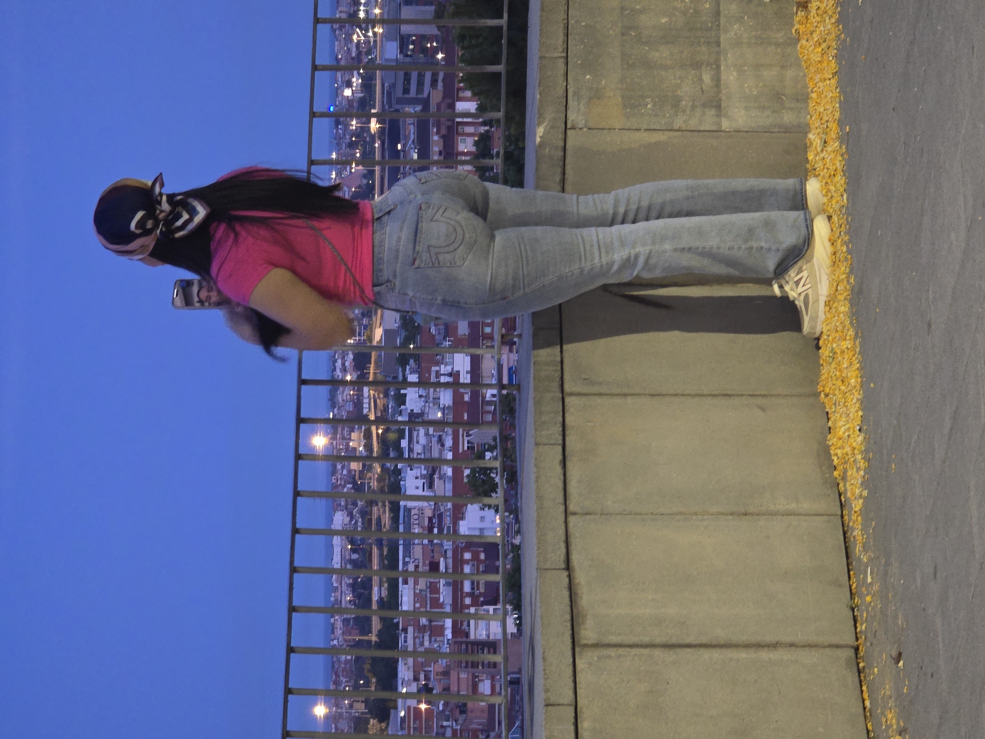 A woman stands on a concrete ledge, facing away from the camera, looking out over a city at dusk. She is wearing a bright pink t-shirt, light blue flared jeans, and light-colored athletic shoes. A patterned scarf is tied around her head. She holds a smartphone in her hands, appearing to take a photograph or video of the cityscape. The city, identified as Cornellà de Llobregat, Spain, is visible in the background with numerous buildings and streetlights illuminated against the deepening blue sky. A metal railing runs along the edge of the ledge in front of her, separating her from the view. The ground in the foreground is asphalt, with scattered yellow leaves or debris.