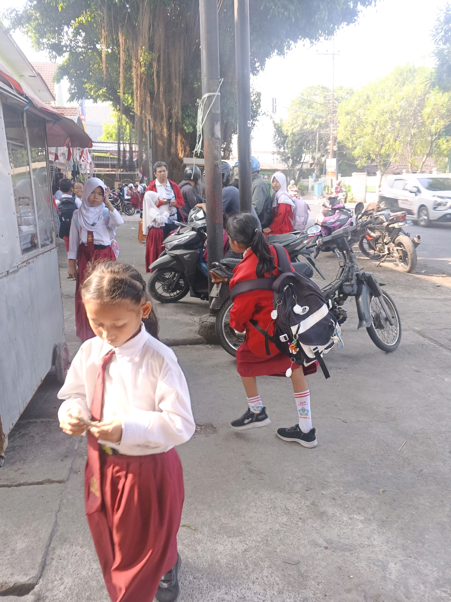 This image depicts a street scene in Krian, Indonesia, likely near a school during morning hours. In the foreground, a young girl in a white shirt and red skirt, with a red tie, is looking down and holding something in her hands. Her hair is pulled back in a ponytail, and she wears white socks with red stripes and black shoes. Behind her, another girl in a similar uniform but with a red jacket is also looking down, her black and white backpack slung over her shoulder. She wears white socks with red stripes and black shoes.
In the background, several other students are visible, some wearing hijabs. A man in a red jacket stands near a stall with awnings. Various motorcycles are parked along the sidewalk, suggesting a busy area. The environment appears to be a public street with a sidewalk and road, lined with trees and buildings. The lighting suggests it is daytime, with the sun casting shadows. The overall impression is of students heading to or from school.