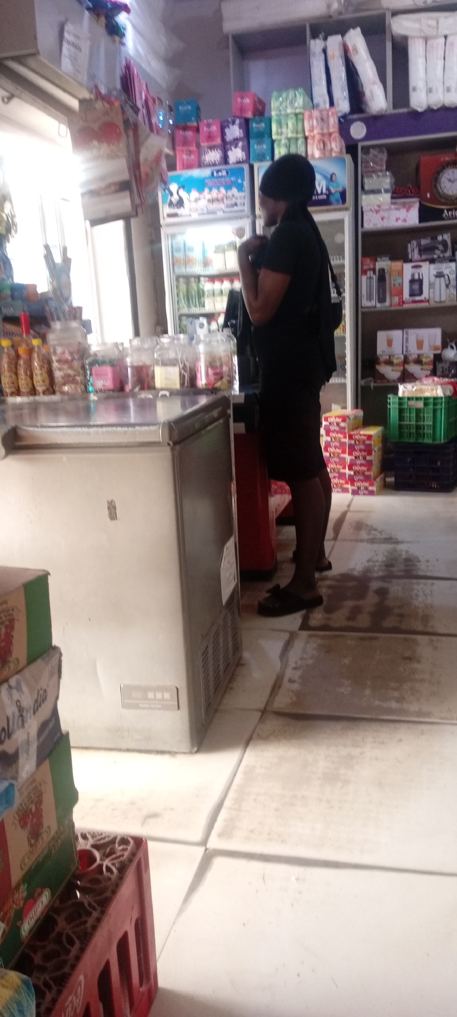 The image shows the interior of a small shop in Fagge D2, Nigeria. A woman, dressed in a black top, black skirt and black sandals, is standing in the shop. She is looking toward a refrigerated display case. To the left of the woman is a large white freezer with jars of snacks on top. Behind the woman, a display case holds bottled drinks and milk. Shelves stocked with various household goods, including paper products, snacks, and small appliances, are visible behind the refrigerated display case. The floor is tiled with light-colored square tiles. Crates of Coca-Cola are stacked near the bottom left corner of the frame.