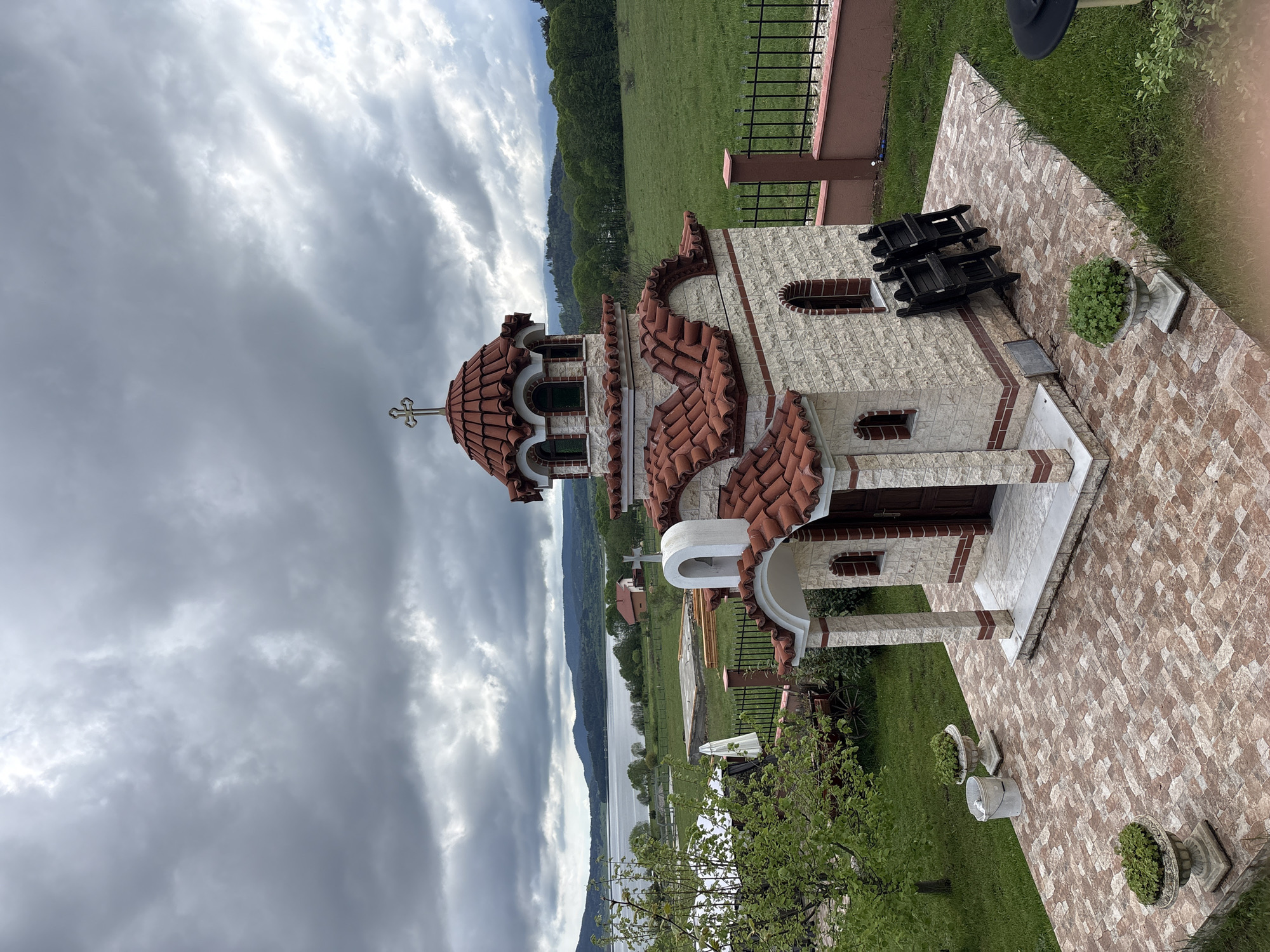 The image depicts a small, stone Orthodox church with a red-tiled roof and a golden cross on its dome. The church is situated in Цигов чарк, Bulgaria. Dark, stormy clouds fill the sky above. In the background, a lake is visible, bordered by forested hills. The church's exterior features brick accents and small, arched windows. In front of the church, there is a paved area with plants in decorative pots, and a small garden. A few wooden chairs are placed near one side of the church. The overall scene suggests a rural setting with a traditional religious building as a central feature.