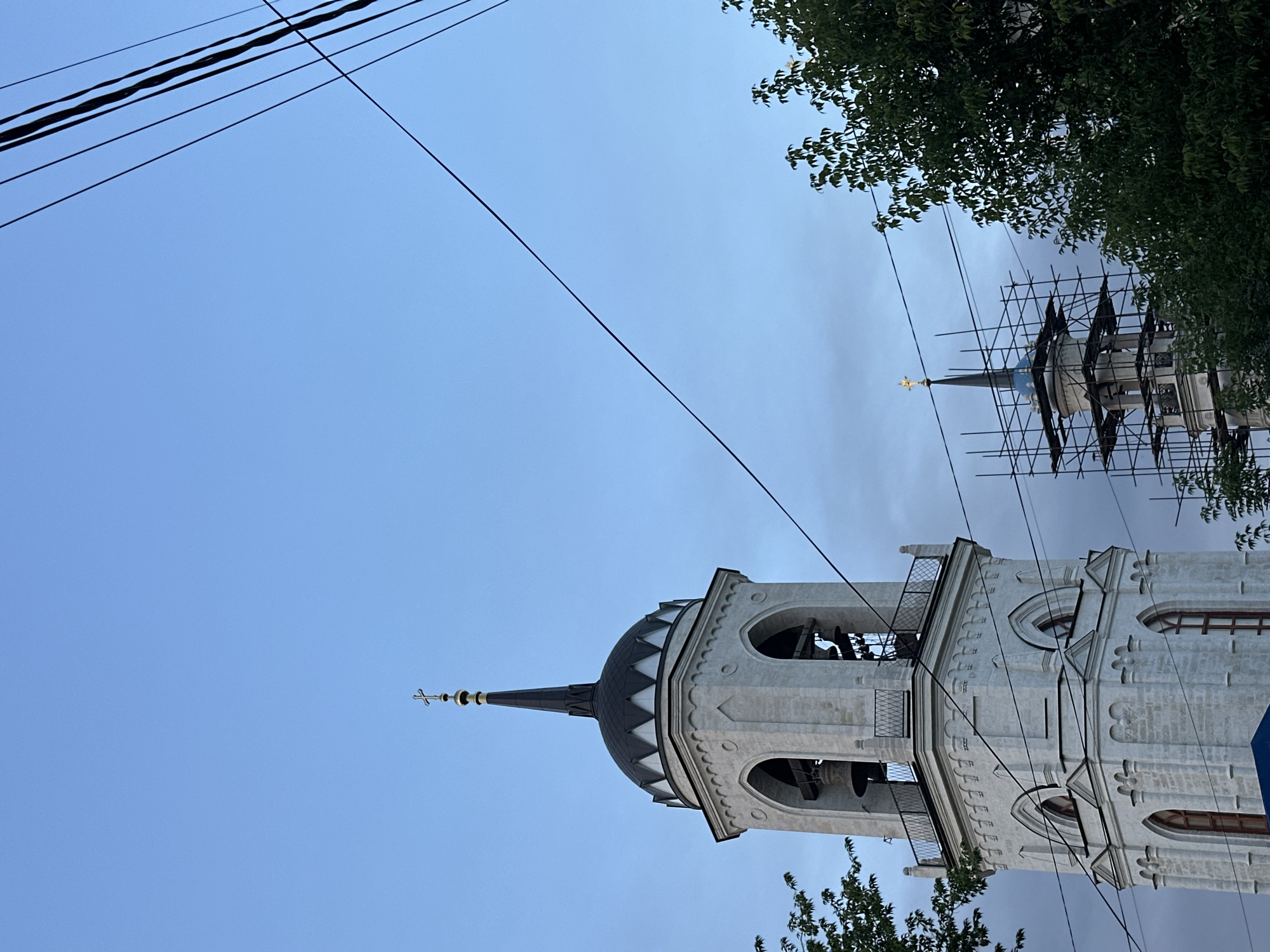 In Быково, Russia, two bell towers of the Church of the Vladimir Icon of the Mother of God are visible against a twilight sky. The foreground features a tall, white bell tower with arched openings, bells, and a black domed spire topped with a gold cross. Electrical wires crisscross the scene, obscuring parts of the towers. To the right, a second, shorter bell tower is partially covered in scaffolding, also topped with a gold cross. Green trees frame the lower edges of the image, and the sky is a gradient of blue and gray clouds.