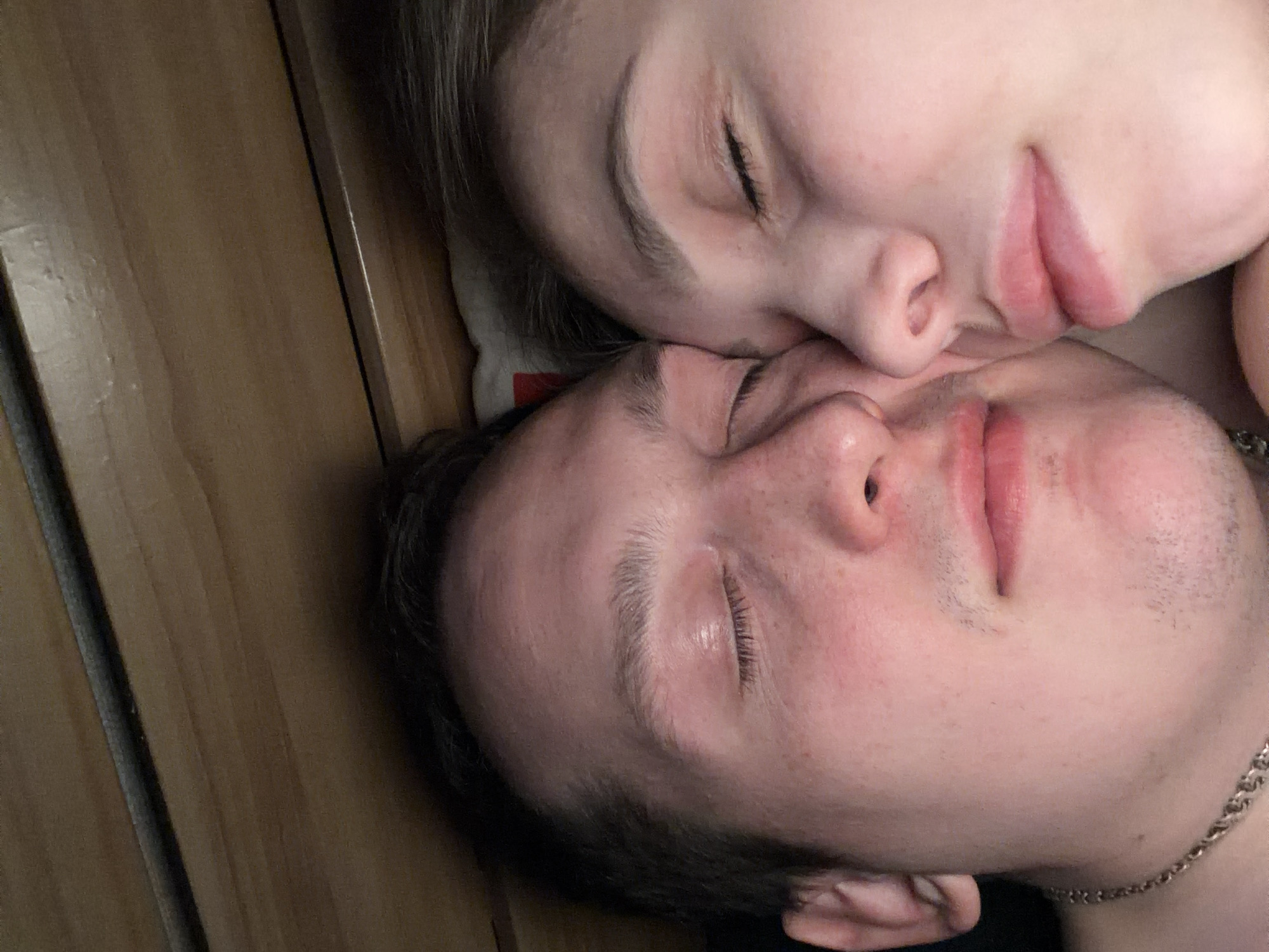 The image shows two people lying down, close together. Both individuals have their eyes closed. The person on top appears to be a light-skinned female, resting her head near the other person's face. The person below is a light-skinned male. A silver-colored necklace is visible on the male's neck. The background consists of a wooden-looking surface. There is no indication of the location being Chaikovsky, Russia, or any specific city.