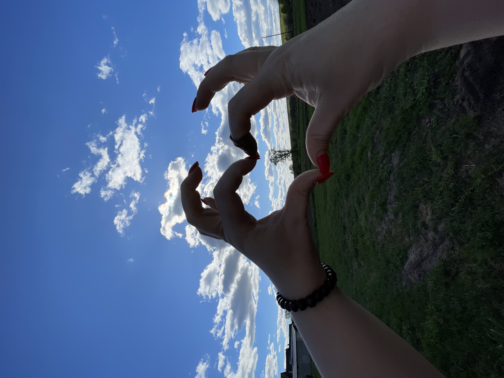 The image shows two hands with red nail polish forming a heart shape against a background of a blue sky with white clouds and a green field. One wrist has a black beaded bracelet. The location is identified as Балдейка, Russia, though there are no specific landmarks in the image to confirm the location beyond the typical rural landscape. A tree is visible in the background on the green field. A section of a low-rise building is visible at the bottom left. The image is taken from a low angle, pointing upwards.