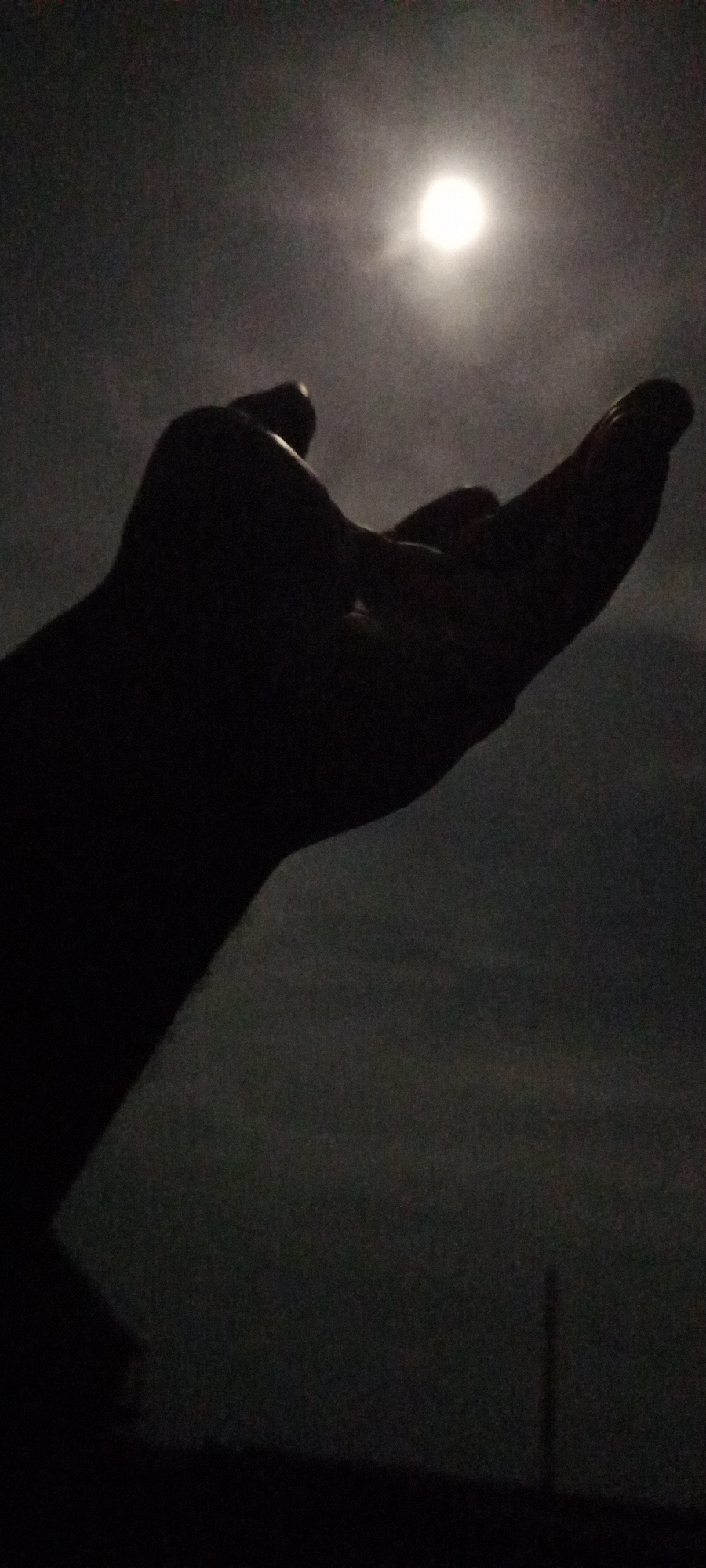 A silhouetted hand is raised towards a bright, round light source, likely the moon, in a dark, night scene. The background suggests an open space, possibly a field or rural area, with a faint outline of a distant structure or pole visible near the bottom. The location is labeled Unknown, Unknown.