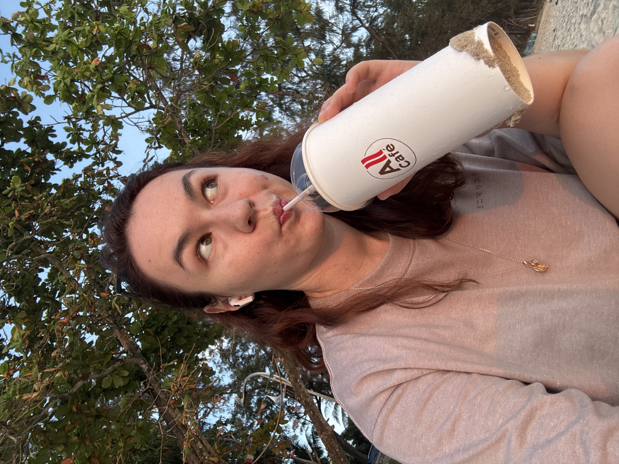 A woman is taking a selfie on a beach in Karon, Thailand. She is wearing a light pink sweatshirt and a gold necklace. She has a white cup with the logo "All Café" in her right hand, and she is drinking from it with a straw. She is also wearing white earbuds. The background shows trees and the ocean. The sun is setting, casting a warm glow on the scene.