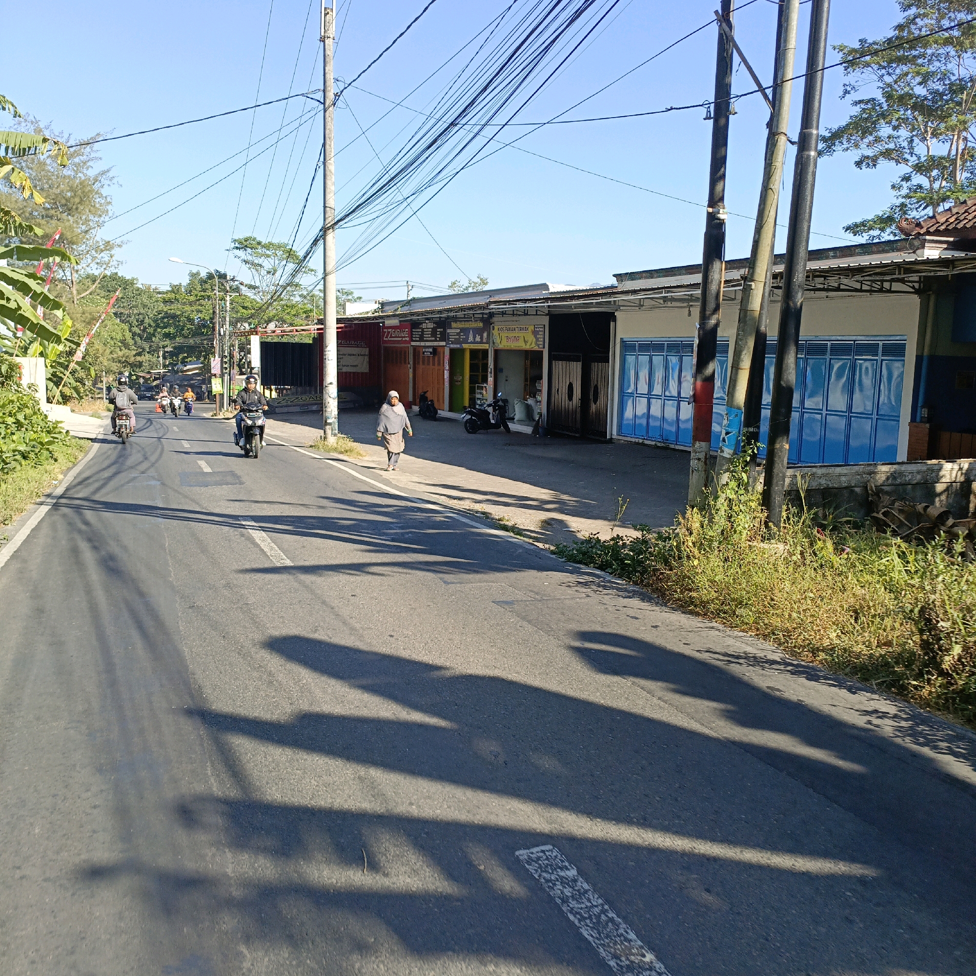 The image shows a street scene in Salatiga, Indonesia, with a clear blue sky and bright sunlight casting long shadows. Several motorcycles are visible on the asphalt road, with some people riding them and one person walking on the sidewalk. Along the side of the road, there are commercial buildings with colorful storefronts and blue-paneled doors. Electrical poles with numerous wires crisscross the scene. The vegetation appears dry and sparse in some areas, with some green trees and banana plants visible. The overall impression is a typical daytime street view in a tropical urban environment.