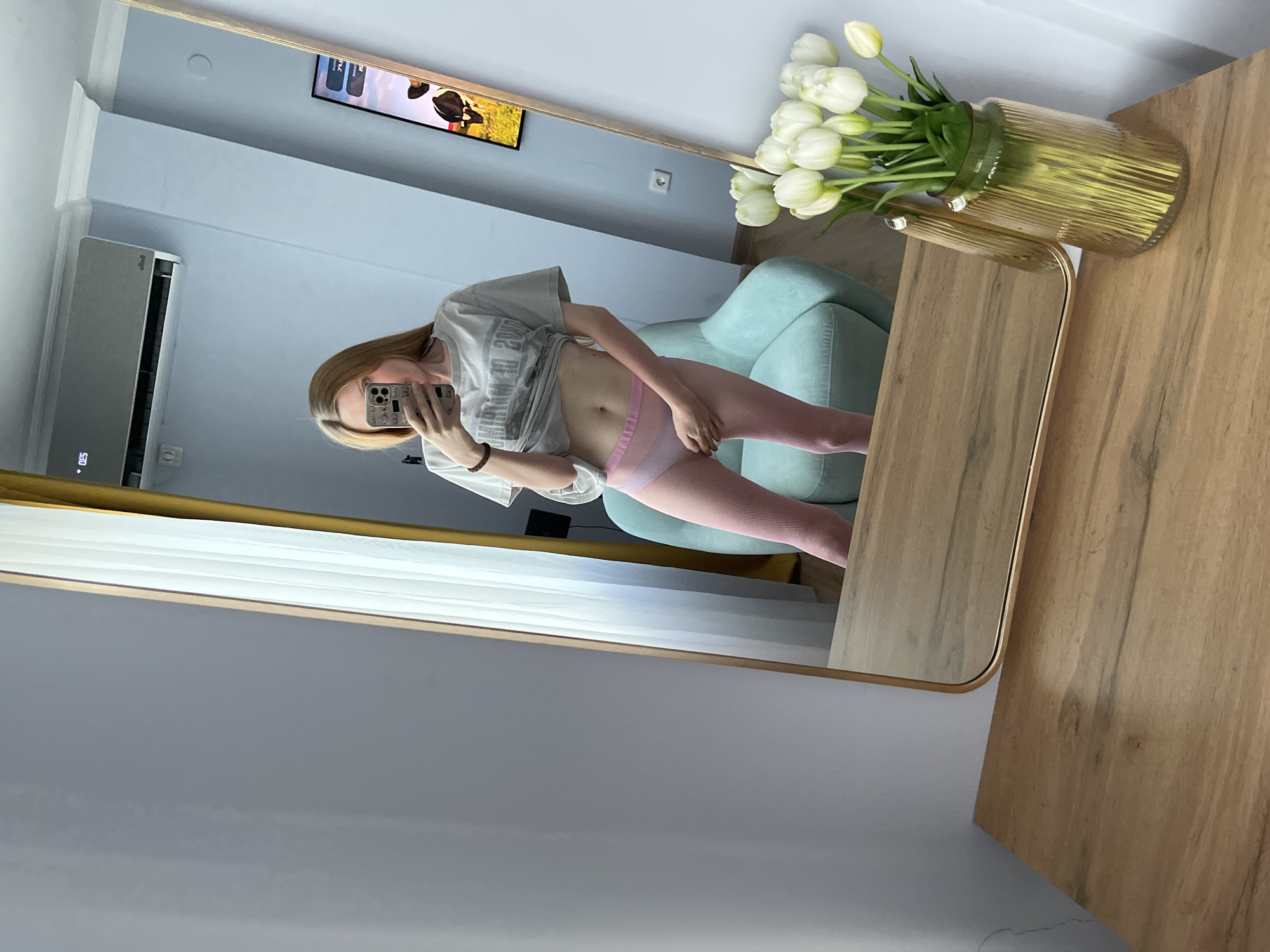 A young woman with blonde hair stands in front of a mirror in what appears to be a bedroom. She is wearing a grey t-shirt and pink textured leggings. The woman is taking a selfie with her phone, which is visible in her left hand. In the reflection of the mirror, a light blue chair and a television displaying an image of cows can be seen. A vase of white tulips sits on a wooden table in the foreground. An air conditioner is mounted on the wall above the woman's head.