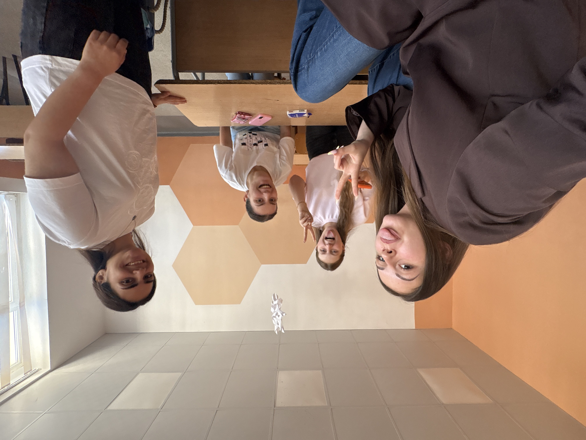 Four young women are in a classroom. One wears a brown blazer and blue jeans. Two others wear white t-shirts and black pants. The fourth wears a white t-shirt and blue jeans. A small white paper object is floating in the air. A table is in the center of the scene with a pink cell phone and a small white box on top. The walls are painted with white and orange hexagonal patterns. The scene is photographed from an unusual rotated angle. The location appears to be a school in Burgas, Bulgaria.