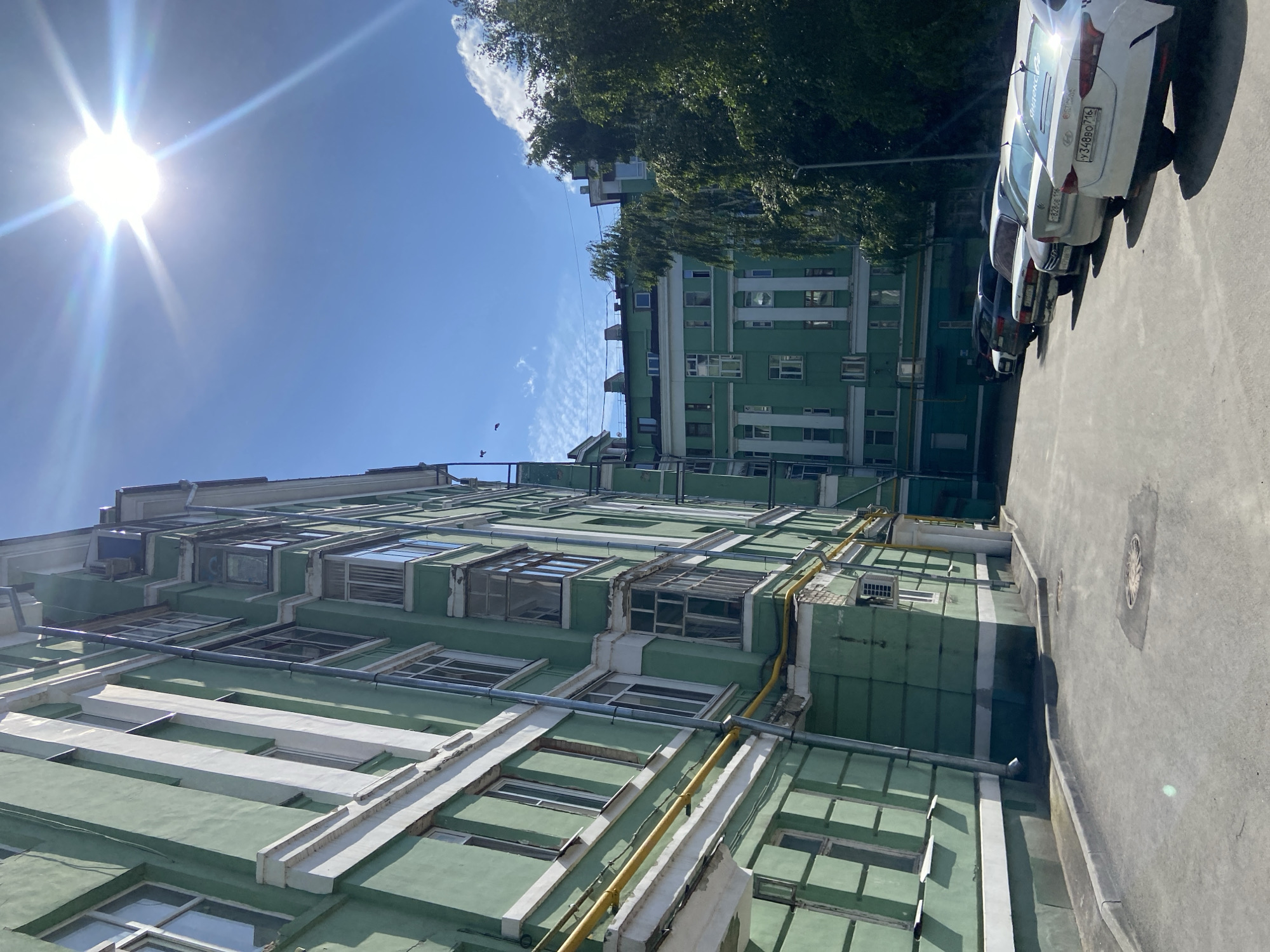 This image depicts a sunlit urban scene in Kazan, Russia, focusing on the exterior of a distinctive light green residential or administrative building with white architectural trim. The bright, high sun casts strong shadows and produces a prominent lens flare in the upper center of the frame against a clear blue sky, which contains a few scattered white clouds towards the upper right.
The primary building, occupying the left and central-left portions of the image, is multi-storied and features numerous windows with white frames, along with several enclosed balconies or bay windows. Visible utility infrastructure includes a yellow gas pipe running horizontally across the building's facade and a silver downspout. The building's exterior shows signs of age, with some peeling paint and weathered sections. In the background, to the right of the main building, another similar green building with white decorative pilasters and windows is partially visible, interspersed with dense green foliage from large trees.
The ground is a paved asphalt surface, likely a courtyard or a side street, with a raised concrete curb running along the base of the buildings. Two circular manhole covers are visible on the pavement. A line of several parked cars, mostly sedans, extends into the mid-ground on the right side. The rearmost car, a white Hyundai, clearly displays a Russian license plate "У348ВО 716", with "716" identifying the Republic of Tatarstan, of which Kazan is the capital. Another vehicle further down the line has a "Yandex" sticker on its rear window. Birds are faintly visible flying in the sky near the upper left quadrant. The overall scene suggests a bright, warm day.