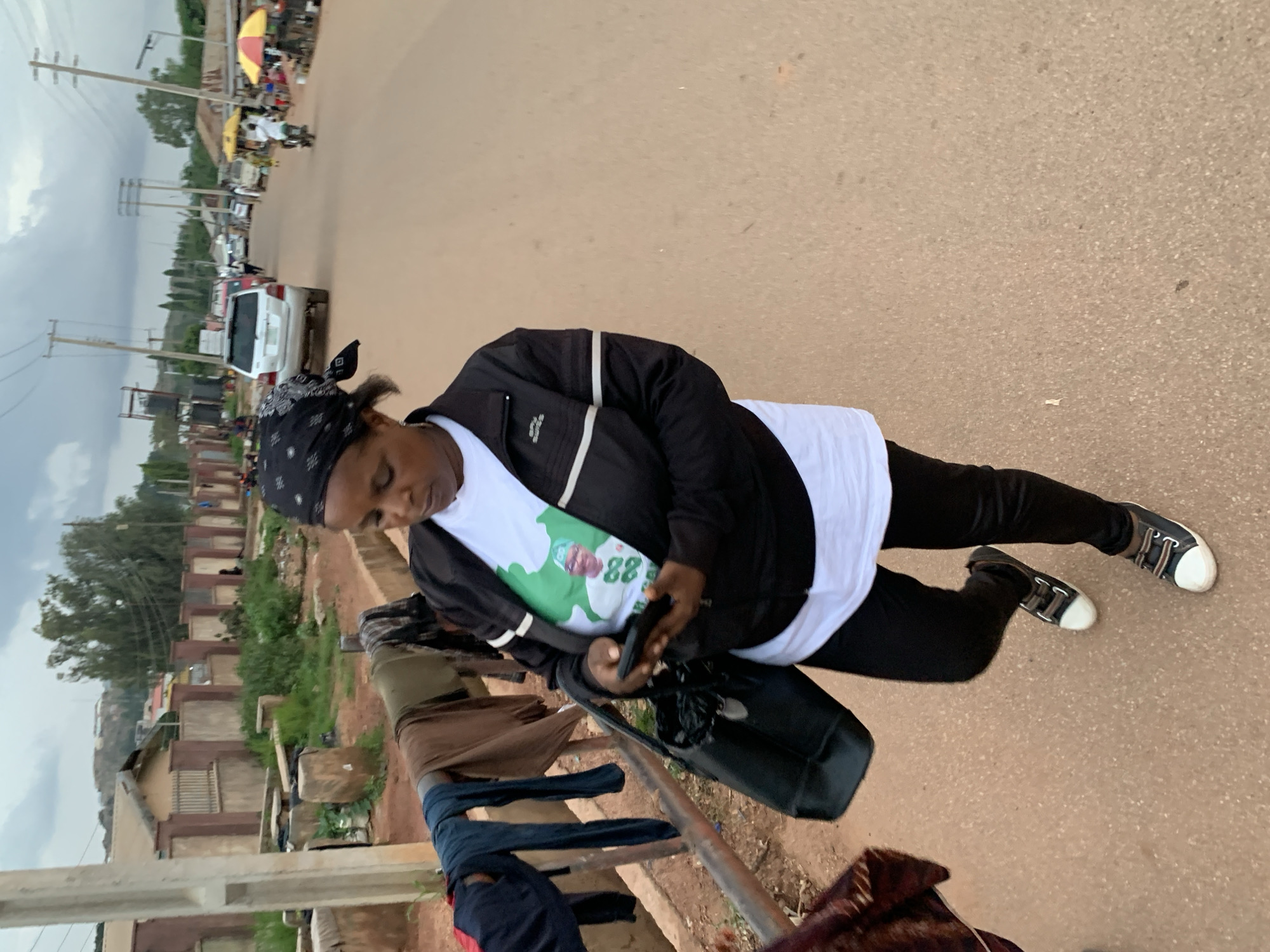 In Gura Top, Nigeria, a woman wearing a black headscarf and a white t-shirt with a political figure printed on it, along with a black jacket and black leggings, walks down a dusty road. She is holding a black smartphone and carrying a black purse. In the background, there are houses with red-tiled roofs and concrete walls, clothing hanging on a railing, several cars, a motorcycle, and market stalls with umbrellas. The sky is overcast.