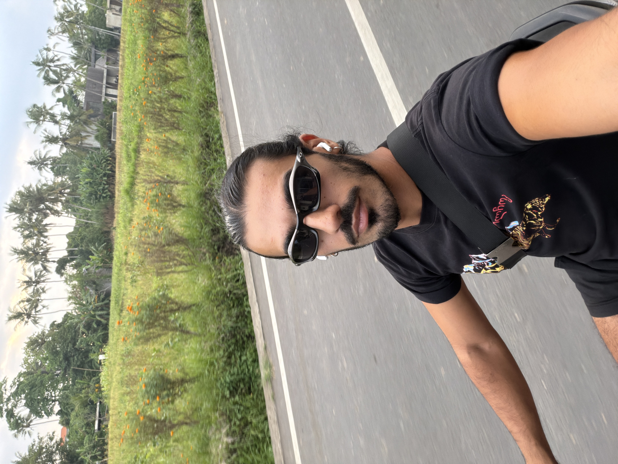 The image displays a close-up portrait of a man, likely in his 20s or 30s, positioned on the right side of a road. He has dark hair pulled back, a neatly trimmed beard and mustache, and is wearing black, sporty sunglasses and a white earbud in his right ear. He is dressed in a black t-shirt that features a colorful graphic, possibly an animal, with partially visible red or pink text that appears to spell "Augustus" in a stylized script. A black strap, likely from a bag or seatbelt, runs across his chest.
The background reveals a gray asphalt road with a distinct white lane marking, running parallel to a lush green field. This field is dotted with small orange flowers and extends towards a dense line of tropical vegetation, including numerous tall palm trees and other leafy plants. Amidst the greenery, several buildings, possibly residential or small structures, are visible in the distance. The sky above is overcast, suggesting a cloudy day or a time when the sun is not directly overhead. The man is looking directly at the camera, possibly taking a selfie while on the move or stopped by the side of the road. The tropical flora and road setting are characteristic of rural or semi-urban areas in Southeast Asia, aligning with the context of Mengwi, Indonesia.