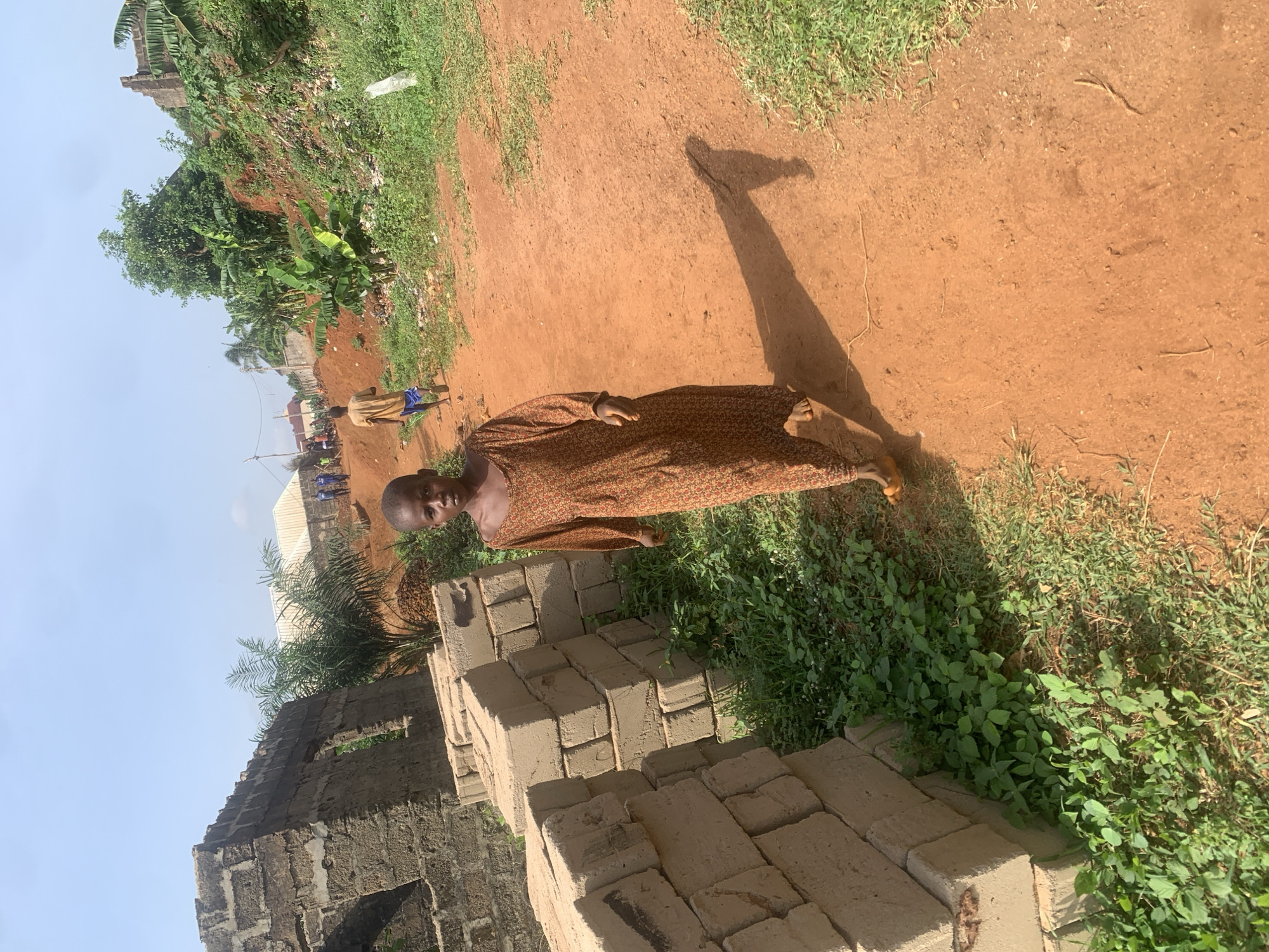 An exterior shot taken at an angle shows a person in a brown, patterned long-sleeved garment standing on a dirt path in Nsugbe, Nigeria. The person's shadow stretches along the path. To the left of the individual is a stack of grey rectangular blocks, possibly bricks or concrete blocks, and an unfinished grey block building in the background. The path is bordered by green vegetation, including grass and leafy plants. In the distance, there are other people in casual clothing on the path near trees and buildings. The sky is partially visible in the upper left corner.