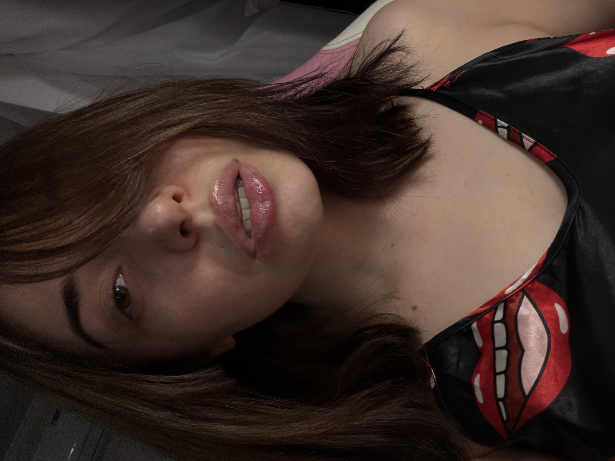 A close-up, waist-up indoor shot shows a young woman with medium brown hair styled in a choppy bob, with bangs partially obscuring her left eye. She is wearing a black satin camisole with a large, stylized red and white cartoon mouth print across the chest. Her lips are glossy and slightly parted, revealing her teeth. The background is dimly lit, with a white curtain visible on the right side and a pink and white object, possibly a chair, also on the right. The lighting is soft and creates a warm glow on her skin.
