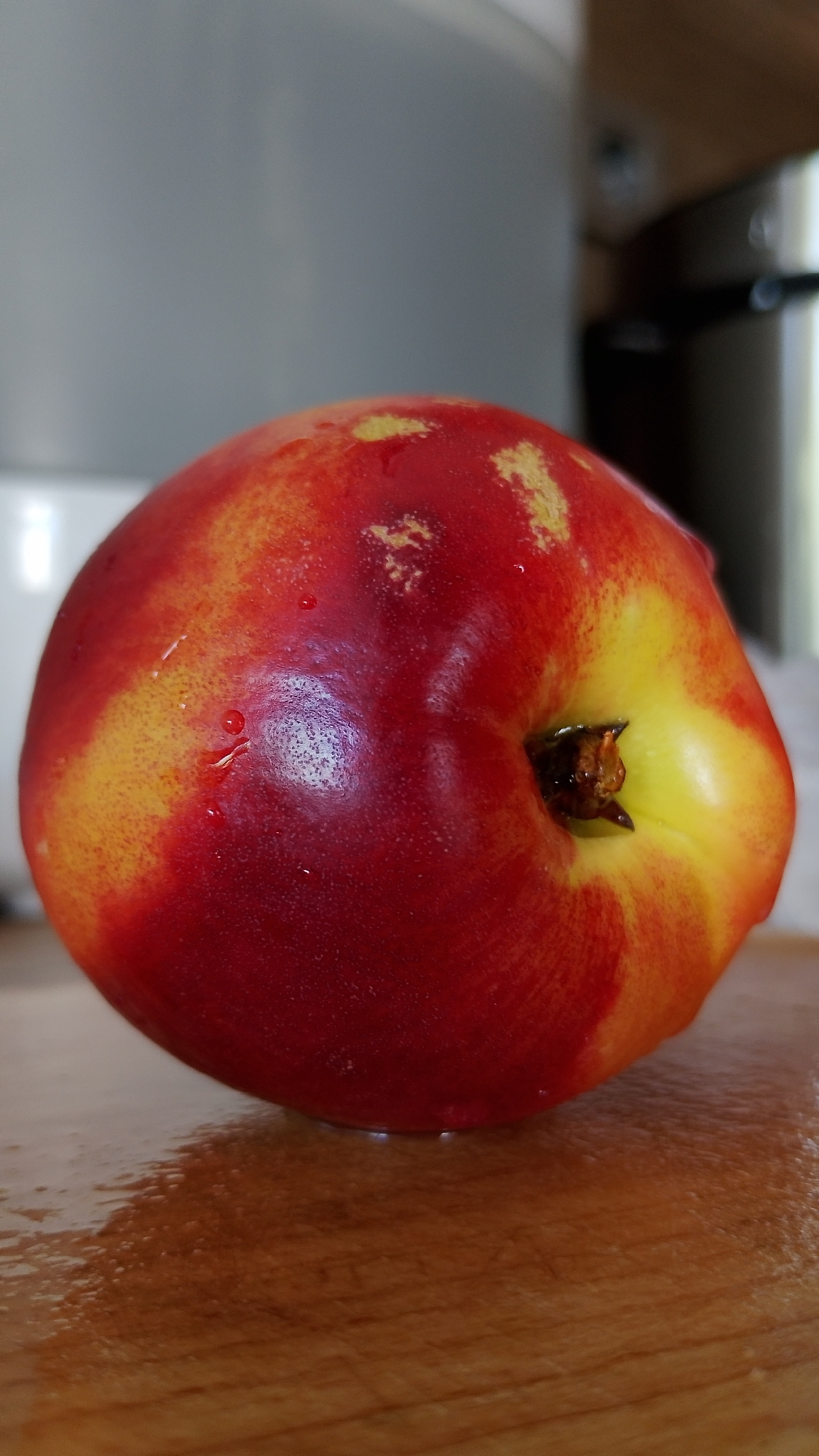A single, ripe nectarine with red and yellow coloring rests on a wet wooden surface. The nectarine is positioned in the center of the frame, with its stem end facing the viewer. Tiny water droplets are visible on the fruit's skin and the wooden surface, suggesting it has recently been washed or is moist. The background is blurred, showing hints of kitchen appliances, but the focus remains on the fruit. No people are present in the image. The location in Smolyan, Bulgaria, is not discernable from the visual information in the image.