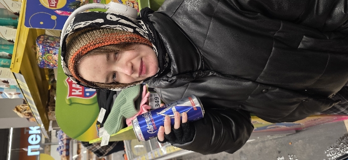 A woman wearing a knit hat, headphones, and a black jacket holds a can of Adrenaline Rush energy drink. She stands near supermarket shelves filled with groceries, including pastries and products from the Lipton tea brand. The scene takes place indoors.