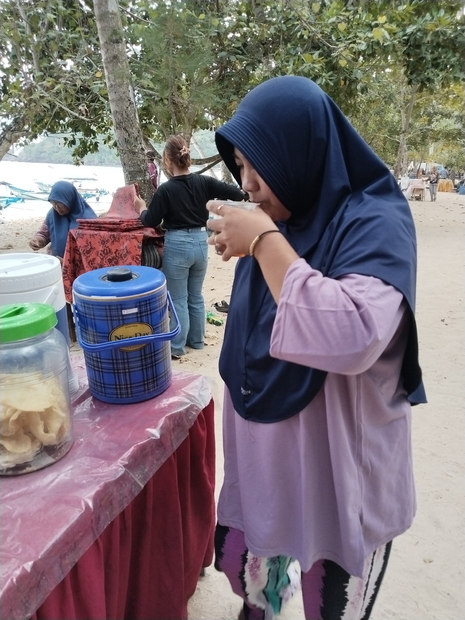A woman in a dark blue hijab and a light purple shirt is drinking from a clear glass. She is standing on a sandy beach near trees. In front of her is a table covered with a red tablecloth and a clear plastic sheet. On the table are a clear jar filled with chips, a blue and black plaid cooler with a "Nice Day" label, and another white container. Behind her, other people are visible, including a woman in a blue hijab and another in a black shirt and jeans. Boats are in the background, suggesting the location is near water, likely the coast. The overall scene depicts a casual outdoor gathering or a vendor stall on a beach. The specific location is identified as Prigi, Indonesia, a coastal area known for its beaches and fishing activities.
