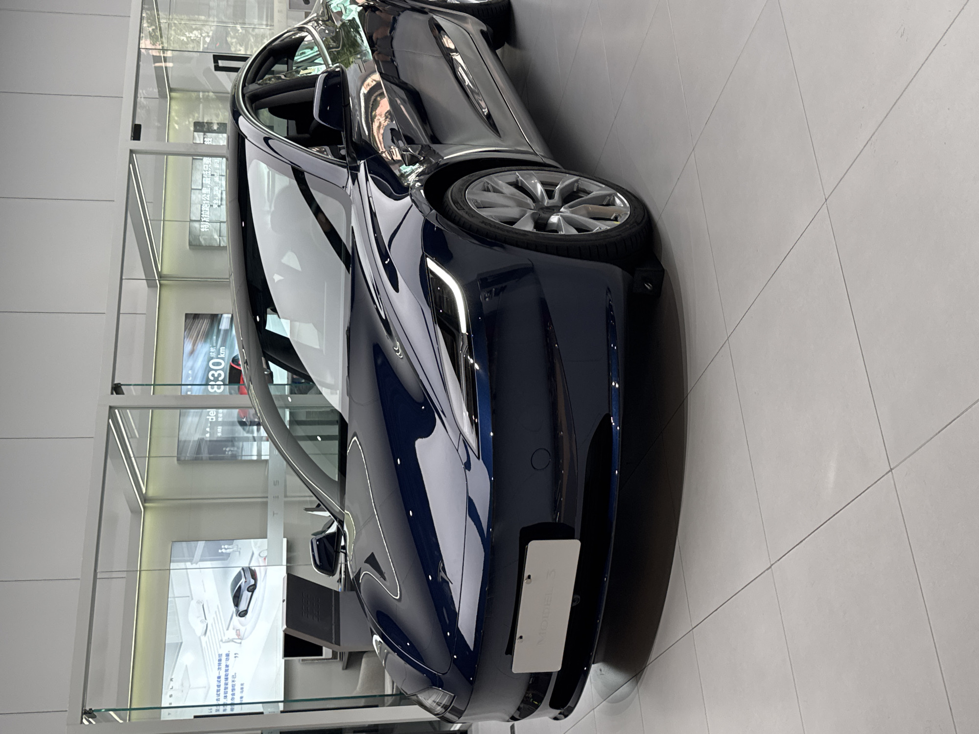 The image depicts a dark blue Tesla Model 3 car in what appears to be a showroom or store. The car is parked on a light grey tiled floor near a glass window. Outside the window, some greenery can be seen, potentially indicating an urban environment. Inside, displays and screens showcase information about Tesla, with Chinese text visible on the displays. The location is identified on the display of 830 km range in Tianhe District, China. The focus is mainly on the car's front, highlighting its design and features.