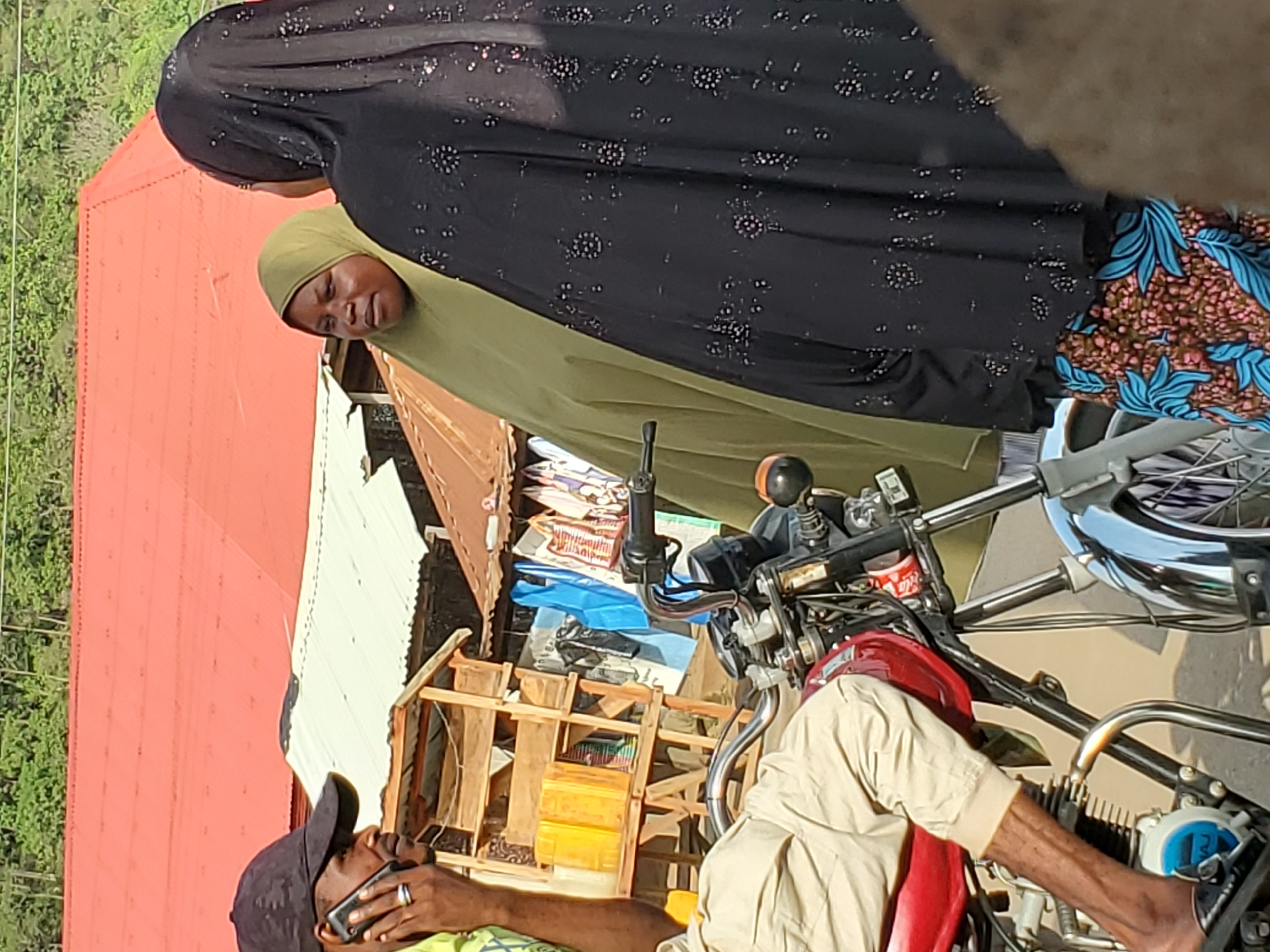 This image depicts an outdoor street or market scene in Mararaba, Nigeria, captured during bright daylight.
In the foreground, a man wearing a black baseball cap and light-colored clothing is seated on a red motorcycle. He holds a phone to his ear, seemingly engaged in a conversation. The motorcycle features a red fuel tank, chrome handlebars, and visible engine components. A small red can, identifiable by the "Coca-Cola" logo, rests near the handlebars. A partial word, "NAJ," is also visible on a blue and white part of the motorcycle's engine.
Standing to the left of the motorcycle are two women. One woman, partially seen from behind, wears a black abaya with sparkling patterns and a green hijab. Next to her, another woman in a green hijab faces the viewer with a somewhat solemn or tired expression.
Behind the people and motorcycle, there's a rustic wooden stall or stand, appearing to hold various items, including blue and yellow plastic containers and stacks of papers. In the background, buildings with prominent reddish-orange and lighter-colored corrugated metal roofs are visible, with lush green trees and foliage in the far distance, suggesting a blend of built environment and natural surroundings. The overall atmosphere indicates a typical, busy daytime scene.