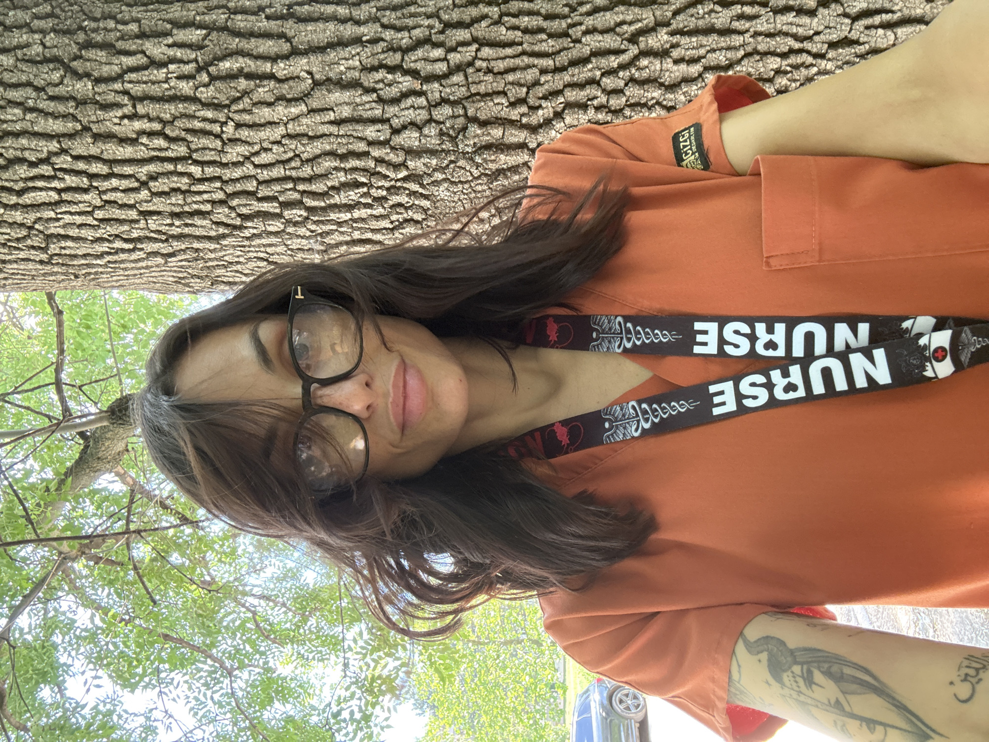 A woman is wearing glasses, an orange uniform, and a "Nurse" lanyard, and is looking at the camera. She has a tattoo on her left arm. Behind her is a tree trunk, green trees, and a car. The image was taken outside. I cannot determine if this is Pleven, Bulgaria.