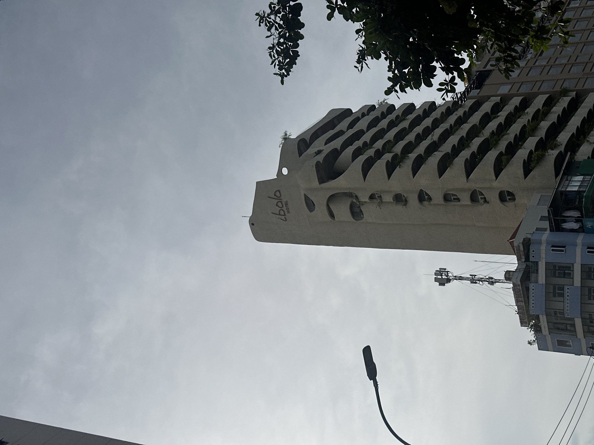 The image is a low-angle, full-shot photograph of the Ibala Hotel in Bac Nha Trang Ward, Vietnam, against an overcast sky. The hotel's facade is a distinctive, light-colored material with a unique architectural design featuring numerous curved balconies, resembling steps or overlapping organic shapes. The word "ibala" is visible in a stylized font at the top of the hotel, with "HOTEL" below it.
To the left of the hotel, a black street lamp with a curved arm extends into the frame. In the foreground, a blue building with multiple windows and a cell tower on its roof is partially visible. Wires are also present in the lower left corner. To the right of the hotel, the dark green foliage of a tree partially obstructs the view of the sky and another building behind it. The overall scene is muted due to the cloudy conditions.