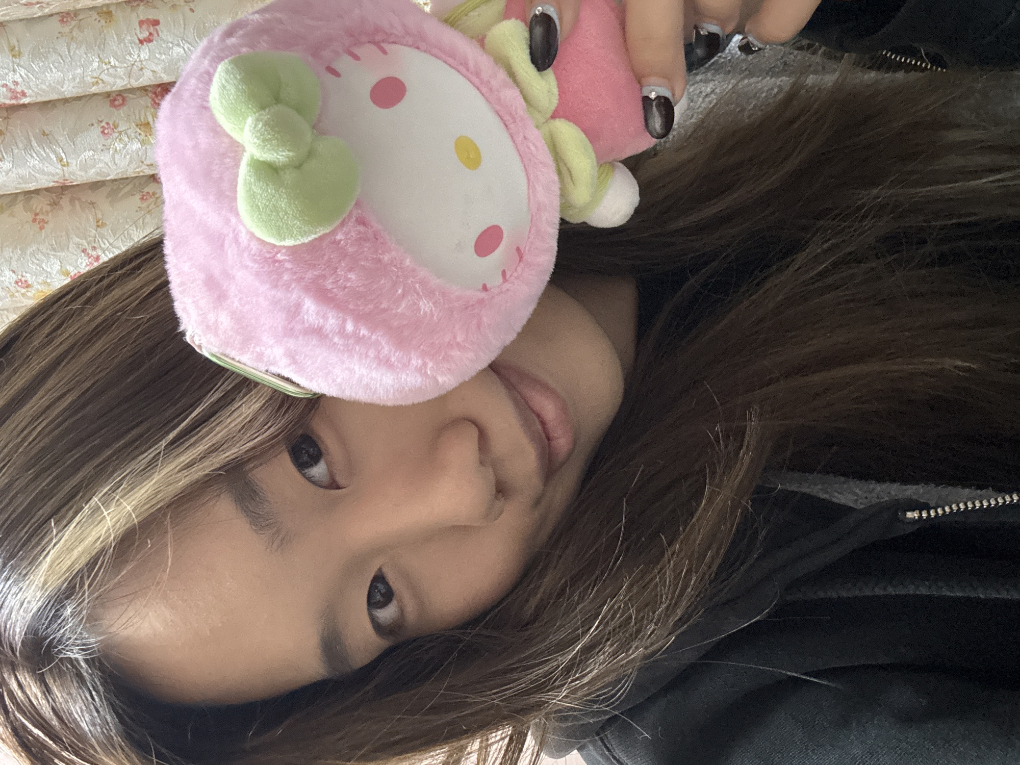 A young person poses for a selfie with a pink, fuzzy Hello Kitty-themed item. The subject's hair is brown with blonde highlights. They are wearing a black hoodie. Their fingernails are painted with a black and silver design. The photo appears to be taken indoors, with floral-patterned bedding visible in the background. There is no verifiable information to suggest the location is in Shapingba District, China.