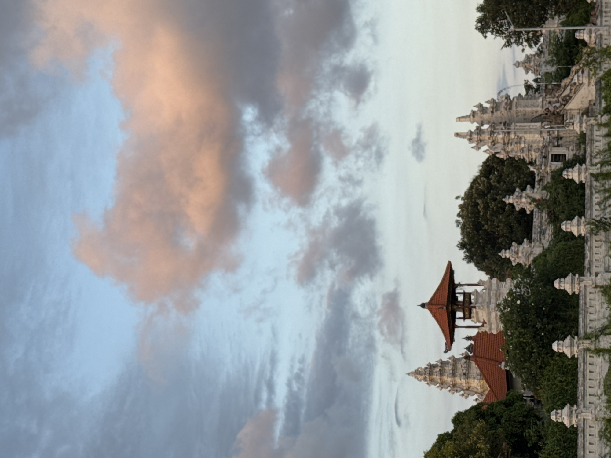 The image shows a Balinese temple complex in Kuta Selatan, Indonesia, under a cloudy sky with patches of light. The architecture is traditional, with layered roofs and ornate white stone carvings. Lush greenery surrounds the buildings.