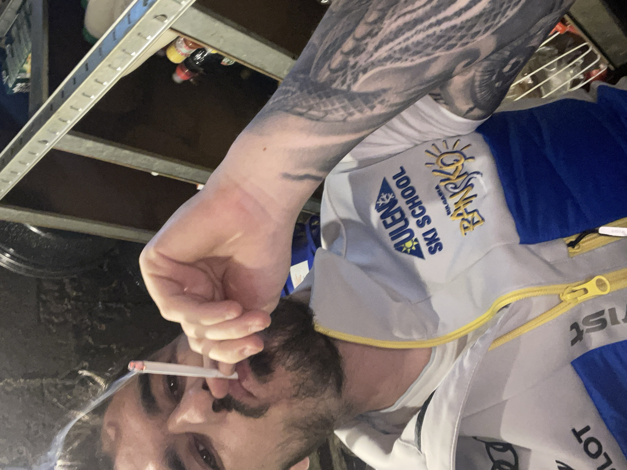 The image captures a close-up, upward-angled shot of a male individual smoking a cigarette indoors. The subject has dark hair and a beard, with his gaze directed towards the viewer. He holds a lit cigarette between his fingers, and a trail of smoke is visible ascending from it.
He is dressed in a white, blue, and yellow jacket, possibly ski wear, featuring a prominent yellow zipper. A white t-shirt is visible beneath the jacket. His right arm is adorned with an extensive, dark-ink sleeve tattoo displaying intricate patterns or designs.
Several texts are visible on his jacket, including "BANSKO" in yellow with a sun icon, and "ULEN SKI SCHOOL" printed below it in blue, suggesting an association with winter sports. Other partially legible texts include "FIS" and "PILOT".
The background is dimly lit and features industrial-style metal shelving units. A few small, colorful bottles or objects are discernible on one of the shelves. The setting appears to be an indoor, possibly utilitarian or storage-like, environment, under low light conditions, likely evening or night. The overall mood is casual.
