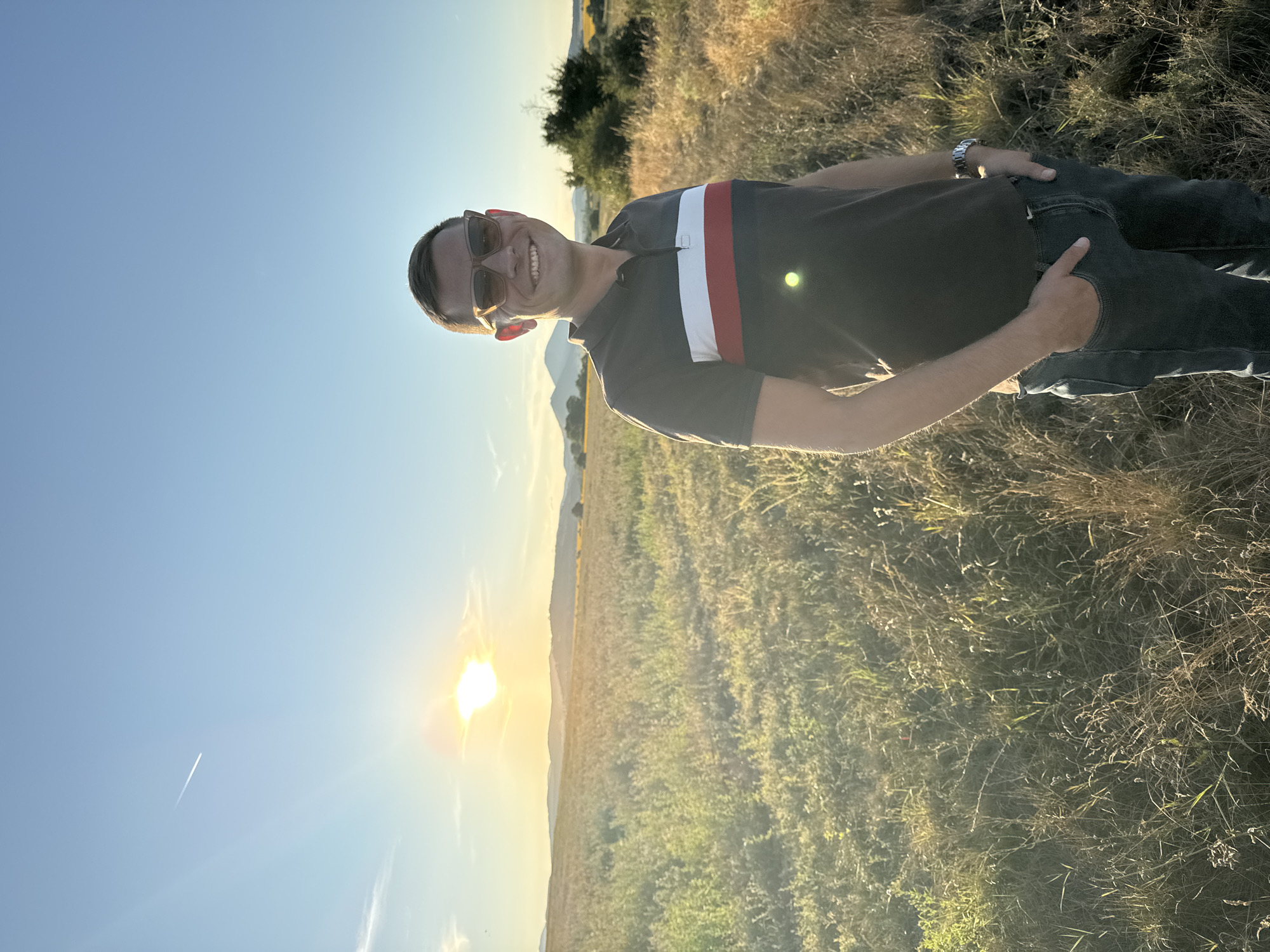 A full-body, outdoor portrait shows a man standing in a field of dry grass and some green vegetation with a hazy sunset in the background. The man, who is looking at the camera, has short brown hair, is wearing sunglasses, and a dark polo shirt with horizontal red, white, and blue stripes across the chest. His hands are in the pockets of his dark denim jeans. The sun is low on the horizon, casting a warm glow over the landscape, and a faint contrail from an airplane is visible in the blue sky. The location is identified as Noevtsi, Bulgaria.