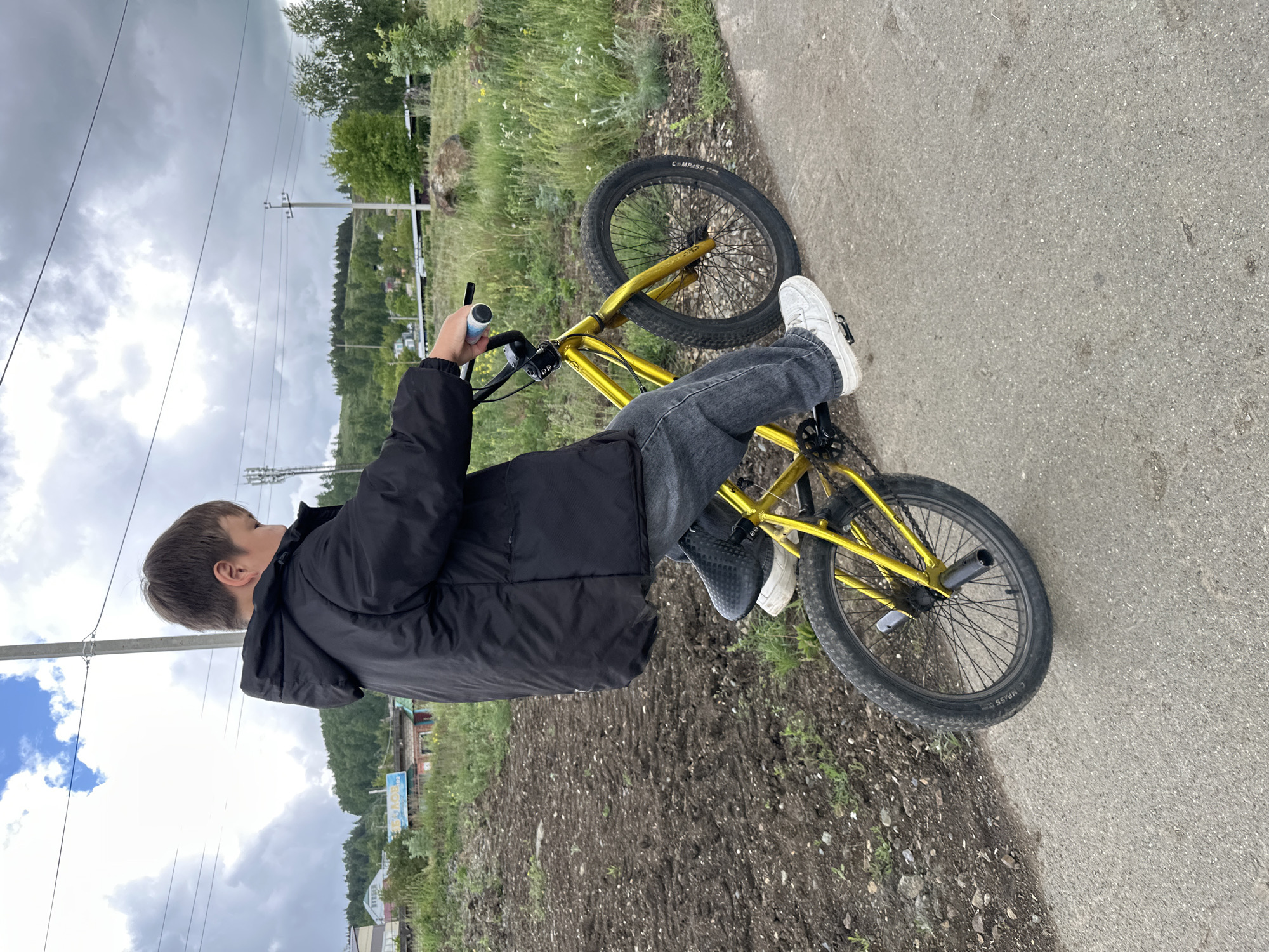 A young person is captured from a slightly elevated, side-rear perspective, seated on a bright yellow BMX bicycle. The image is rotated 90 degrees counter-clockwise, showing the scene on its side. The individual, a young male with short, light brown hair, wears a black puffy jacket, dark grey jeans, a white sneaker on his right foot, and a black sneaker on his left. He is holding the bicycle's handlebars, seemingly paused on a paved surface next to a dirt and grass strip.
The bicycle features a striking yellow frame and handlebars, with black tires, seat, and pedals. The word "COMPASS" is visible on the sidewall of the front tire.
The setting appears to be a semi-rural or exurban area. To the right of the paved path, the terrain transitions into a dirt patch with sparse greenery. In the midground and background, rolling hills densely covered with green trees extend into the distance. A few scattered buildings are visible among the trees on the lower slopes. Multiple power lines and utility poles span across the scene horizontally. The sky above is largely overcast with grey clouds, though hints of brighter sky and blue patches are visible. The overall lighting suggests it is daytime, under cloudy conditions. No strong emotions are discernible from the person's posture or visible features.