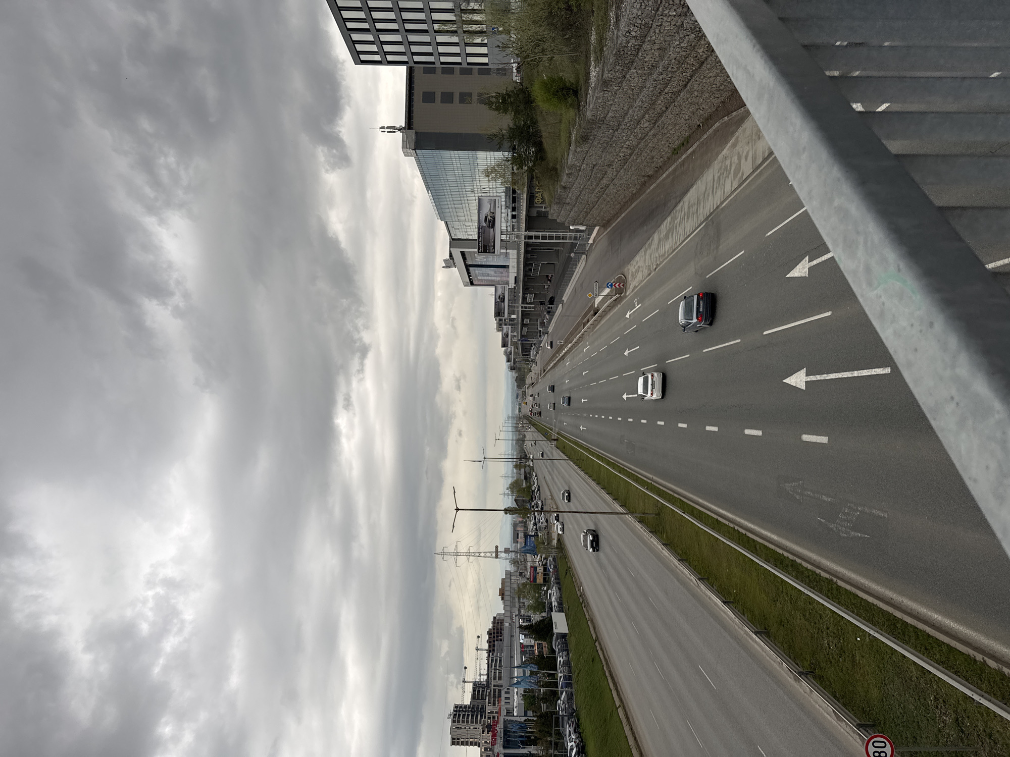 The image captures a wide, multi-lane highway or major road viewed from an elevated perspective, likely a pedestrian overpass. The road stretches into the distance under a uniformly overcast sky.
The highway features multiple lanes of traffic flowing in both directions, separated by a grassy median. White dashed lines mark the individual lanes, and large white arrows are painted on the asphalt, indicating directions such as straight, left turn, and a combination of straight/right turn. Several cars are present on the road, with some appearing to move in both directions, and a higher density of traffic is visible further down the road.
To the left of the highway, a series of modern buildings line the thoroughfare. Some structures feature large glass facades, while others display grid-like window patterns. Large billboards and digital screens are affixed to these buildings; one shows a car advertisement, and another has the brand "OPEL" faintly visible. An elevated gabion wall (stone-filled cages) runs alongside the road, bordering an embankment. Further in the distance on the left, construction sites with cranes indicate ongoing development, and numerous cars are parked in lots adjacent to the buildings.
The right foreground of the image is partially framed by the metal railing of the overpass. Below this railing, a circular red-bordered sign with the number "80" in white on a black background is visible, indicating a speed limit of 80 (likely km/h). The weather is cloudy with a grey, diffused light suggesting daytime. Power lines and streetlights are also notable elements along the roadside. No distinct people are visible in the scene. The overall setting is an urban or suburban environment in Sofia, Bulgaria.