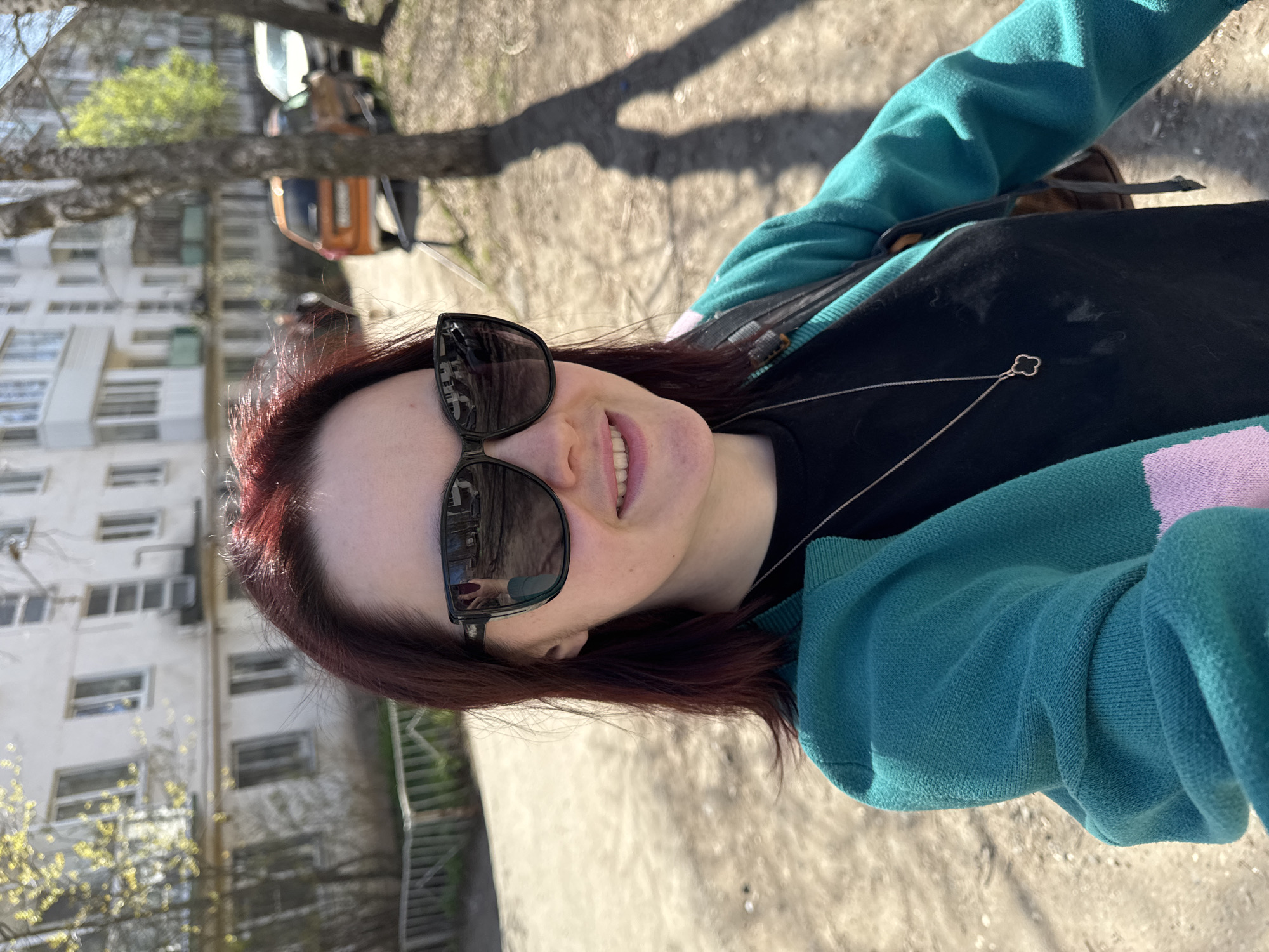 A woman with dark red hair and wearing sunglasses is taking a selfie outdoors. She is wearing a black shirt and a turquoise and lavender cardigan. A silver necklace with a clover pendant is visible. The background shows an apartment building, trees, and a paved area with shadows. The reflection in the sunglasses suggests the presence of another person. The location is Izhevsk, Russia.