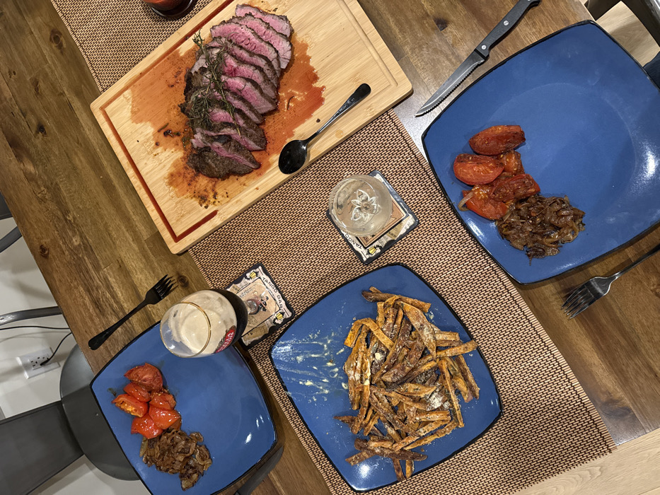 The image displays a dining table prepared for a meal, viewed from an overhead perspective. The table surface is a light-brown wood grain, partially covered by two woven brown placemats.
Central to the scene is a wooden cutting board, approximately rectangular with rounded corners and a perimeter juice groove, containing a sliced steak. The steak exhibits a medium-rare doneness, with pink interior flesh and a seared exterior, garnished with multiple sprigs of rosemary. Reddish-brown liquid, likely meat juices, is pooled within the cutting board's groove and beneath the steak. A silver serving spoon rests on the cutting board to the left of the steak.
Three square blue plates with rounded edges are arranged on the table.
The plate in the upper left, positioned on a placemat, holds a portion of roasted red tomatoes and caramelized brown onions. A black metal fork is placed beside this plate.
The plate in the middle-left, also on a placemat, contains a pile of sweet potato fries, dusted with a white substance consistent with cheese or seasoning. Visible crumbs and residue suggest partial consumption.
The plate in the lower right, off the placemat, holds an identical portion of roasted red tomatoes and caramelized brown onions. A black-handled steak knife and a black metal fork are positioned to the right of this plate.
Two beverages are present. A stemmed glass containing a dark beer with a head is situated on a square coaster bearing the text "TANGO BAR" next to the upper-left plate. A clear, short glass containing a clear liquid (likely water) is also on a "TANGO BAR" coaster, positioned between the middle-left and lower-right plates.
In the upper-left background, a dark grey chair back is partially visible. Behind the chair, a light-colored wall with a white electrical outlet is visible.
There are no individuals present in the frame. The scene is indoors, likely a domestic dining area. The image provides no specific visual cues to determine a city location within Los Angeles, United States of America.