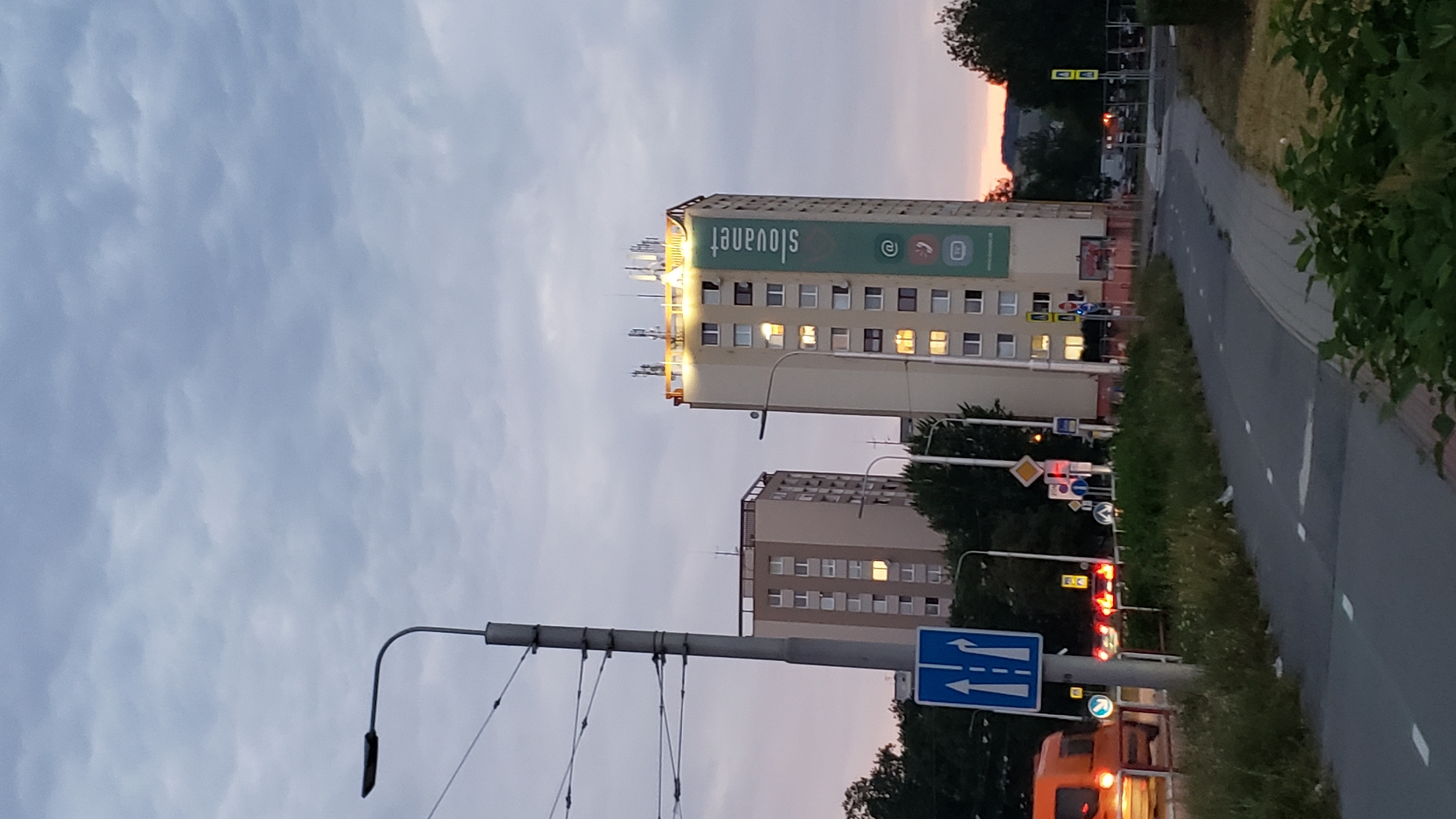 The image shows a street scene in the city of Prešov, Slovakia, at dusk. In the foreground, there is a paved path with white markings, likely a sidewalk or bike lane, bordered by grass and some greenery. A street light with a curved arm extends into the upper left of the frame, attached to a tall, segmented metal pole.
Dominating the mid-ground are two tall residential buildings. The building on the left is lighter in color, with evenly spaced windows. The building on the right is also a light color, taller, and features a large green banner on its side with white text and icons. The text reads "slovaknet" vertically, with an "@" symbol, a phone icon, and a chat bubble icon below it. The upper edge of this building is illuminated by bright lights. Both buildings have multiple antennas on their roofs.
In the background, trees and more residential buildings are visible. The sky is overcast with dramatic, textured clouds, tinged with the colors of a setting sun, suggesting a sunset or sunrise. Several traffic signs are present, including a blue directional sign with white arrows, and a diamond-shaped yellow sign with a red border. An orange vehicle is partially visible on the left side of the image, its headlights and taillights illuminated. The overall atmosphere is that of an urban residential area during twilight.