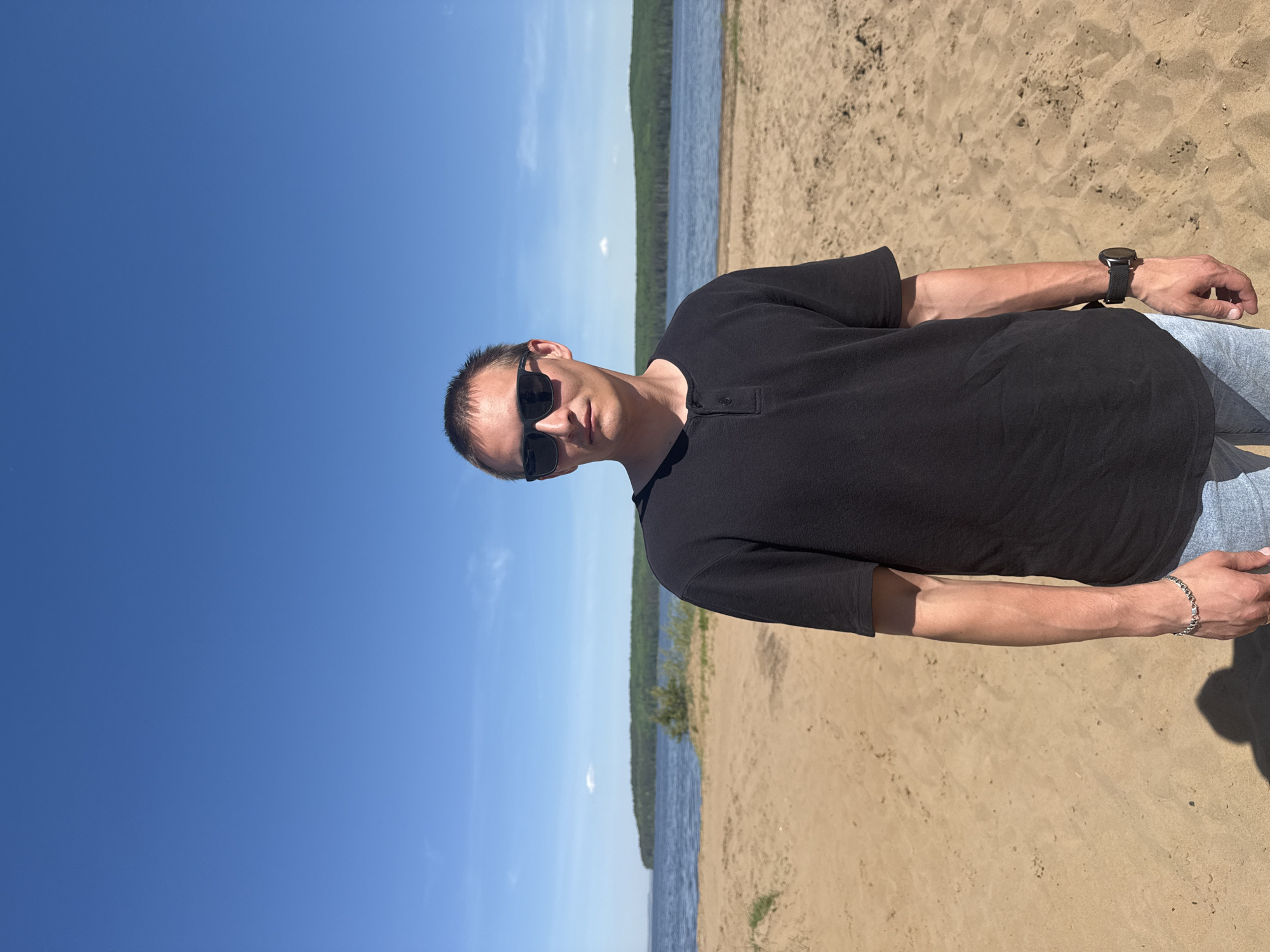 A man wearing sunglasses and a black short-sleeved button-up shirt stands on a sandy beach. He is wearing light-wash denim jeans and a watch on his left wrist. He also has a bracelet on his right wrist. Behind him, there is a body of water bordered by a grassy shoreline. The sky is clear and blue. The location is likely a beach near the city of Chaikovsky, Russia, based on the user's instructions.