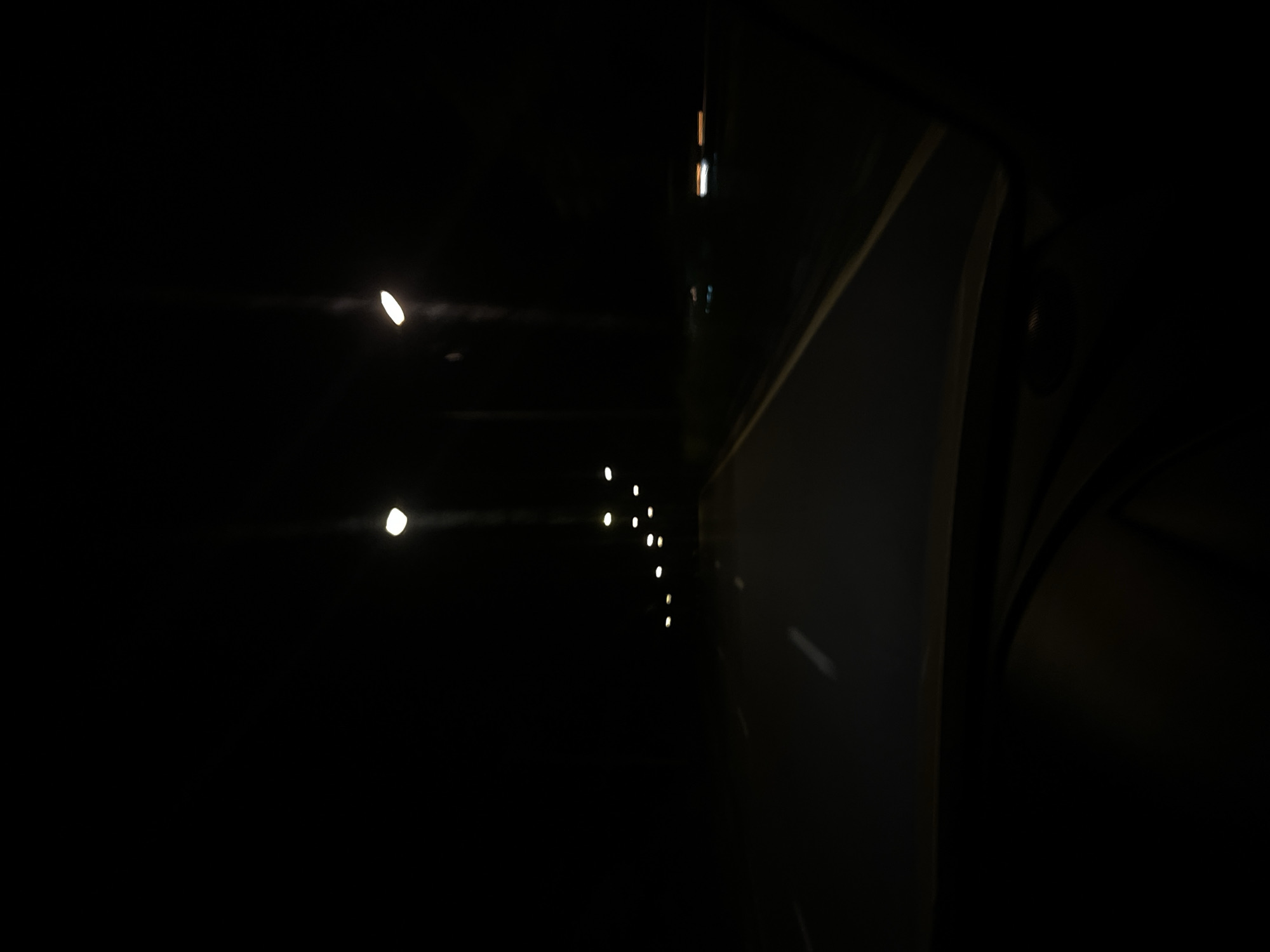 A nighttime, low-light, point-of-view shot from inside a vehicle driving on a dark road. The road appears to be an asphalt highway with lane markings visible. Streetlights line the right side of the road, casting pools of light. The lights are spaced out and appear to be overhead lamps on tall poles. The sky is completely dark, indicating it is nighttime. The foreground shows the interior of a car, possibly a dashboard or windshield, which is also dark. There are no visible people or specific landmarks in the image, making it impossible to determine the exact location within Wichit, Thailand. The image has a slightly blurred or out-of-focus quality due to the low light conditions and movement.