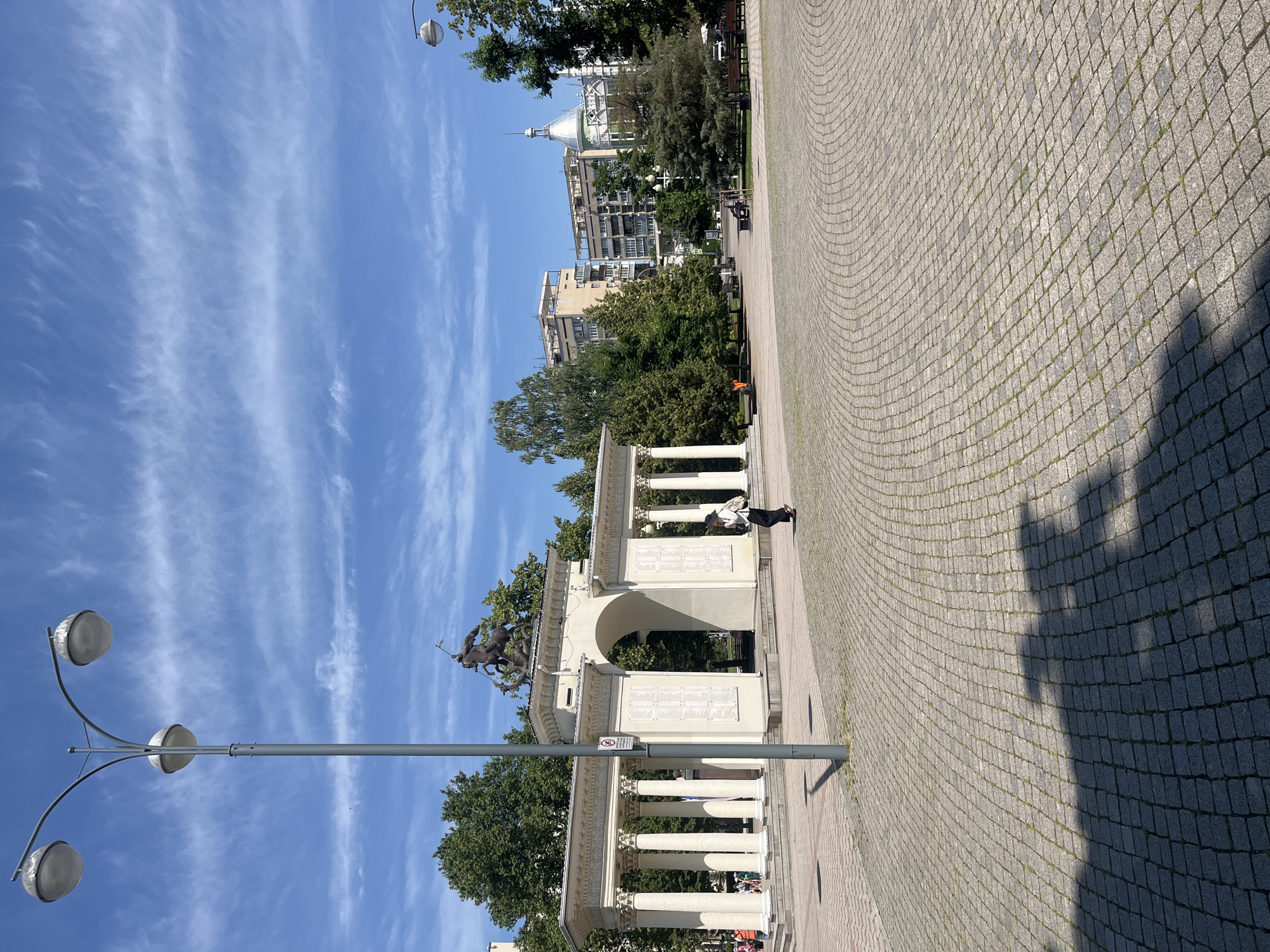 The image shows a view of the Victory Arch in Krasnodar, Russia. The arch is a white, neoclassical structure with columns and an attic. A bronze sculpture of a warrior on horseback is mounted on top of the arch. In front of the arch is a large, paved square. A lamppost with three globes stands in the foreground. In the background, there are trees and buildings. A woman in a white shirt and black pants walks across the square. The sky is blue with some wispy clouds.