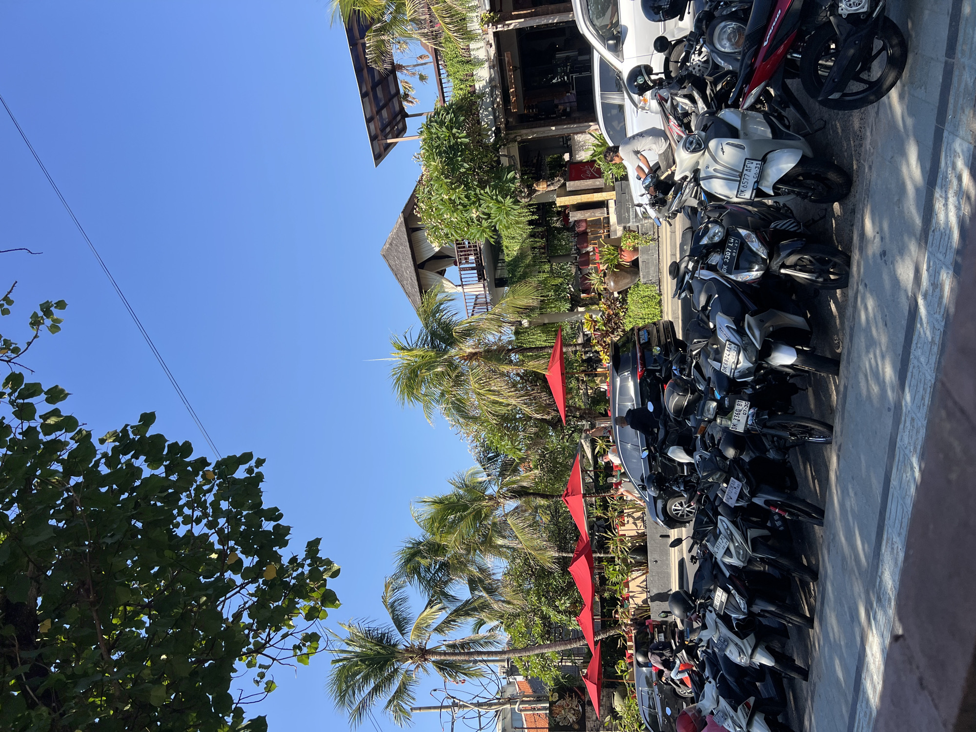 In Kuta, Indonesia, a row of parked motorcycles lines a paved street in front of a multi-story building with a thatched roof. Palm trees and lush green foliage surround the building, which features outdoor seating with red umbrellas. Several cars and motorcycles are visible on the street, and people are seen sitting at tables and riding motorcycles. The sky is clear and blue, and the sun is shining.