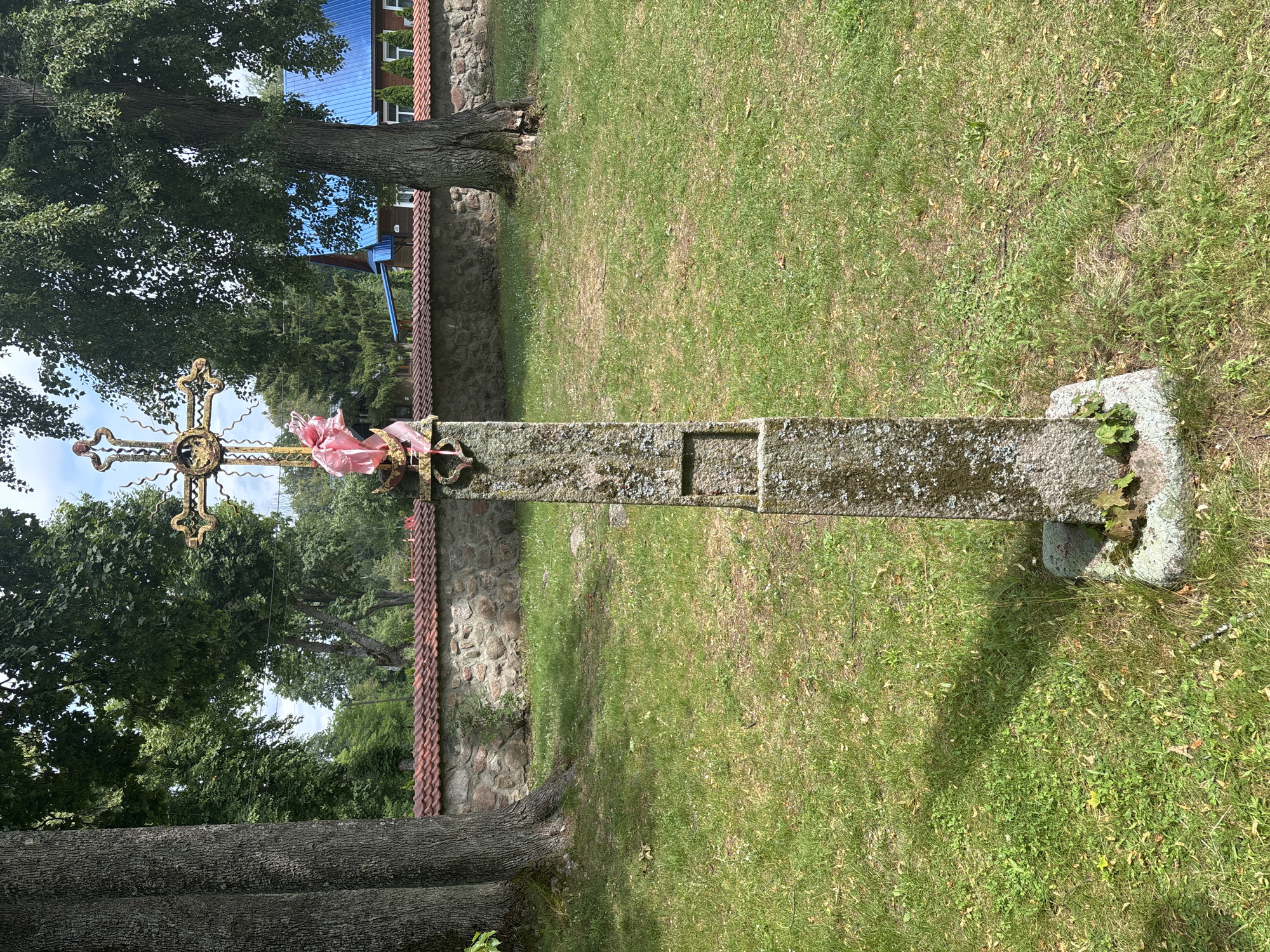 The image depicts a weathered stone monument with a decorative metal cross at its apex, situated in an outdoor setting that appears to be a cemetery or churchyard in Puchły, Poland. The stone base of the monument is rough-hewn and moss-covered, with a visible square indentation on its front. A bright pink ribbon is tied around the cross, suggesting a recent adornment.
Behind the monument, a low stone wall with a terracotta tile roof runs horizontally across the frame. Beyond the wall, a building with a bright blue roof is visible, likely a church or another structure associated with the location. Large trees with lush green foliage frame the scene, with their trunks on the left and right sides of the image. The ground is covered in green grass, interspersed with fallen leaves and small wildflowers. The sky above is partly cloudy, with patches of blue peeking through. The overall impression is one of a tranquil, perhaps historical, rural site.