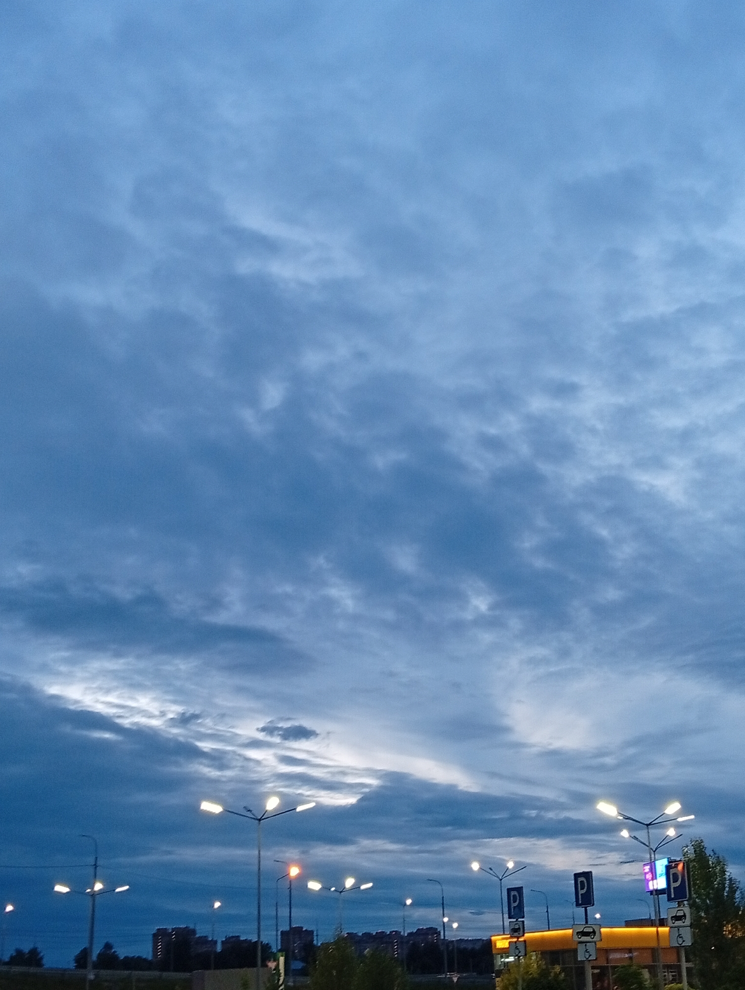 The image depicts a cloudy sky over a city landscape, likely taken during twilight hours. Several streetlights are illuminated, casting a warm glow against the darkening blue and gray clouds. In the background, buildings and trees are visible. The scene appears to be from Pushkino, Russia, as indicated by the presence of parking signs with the letter "P" and symbols indicating parking for people with disabilities. The overall impression is one of an urban environment at dusk, with artificial lights beginning to dominate the fading natural light.
