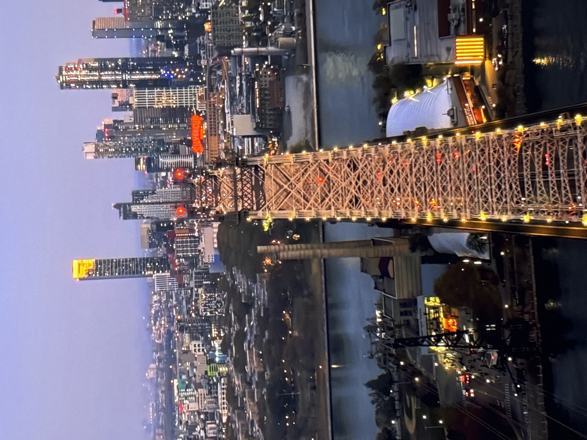 An aerial view captures a bustling city skyline at dusk, dominated by a large suspension bridge illuminated with numerous lights. The bridge stretches across a body of water, separating the viewer from the cityscape. Tall buildings, some with glowing windows and illuminated signage, form a dense urban backdrop. On the bridge itself, vehicle traffic is visible, with red taillights indicating movement. The sky above is a soft gradient of purple and grey, suggesting the transition from day to night.
The image does not depict Burgas, Bulgaria, but rather a city with a prominent bridge and a dense urban environment. The architecture and style of the buildings suggest a North American city. There are no people or specific actions clearly discernible in the image beyond general city life. The environment is urban, featuring water, bridges, buildings, and a cloudy sky.